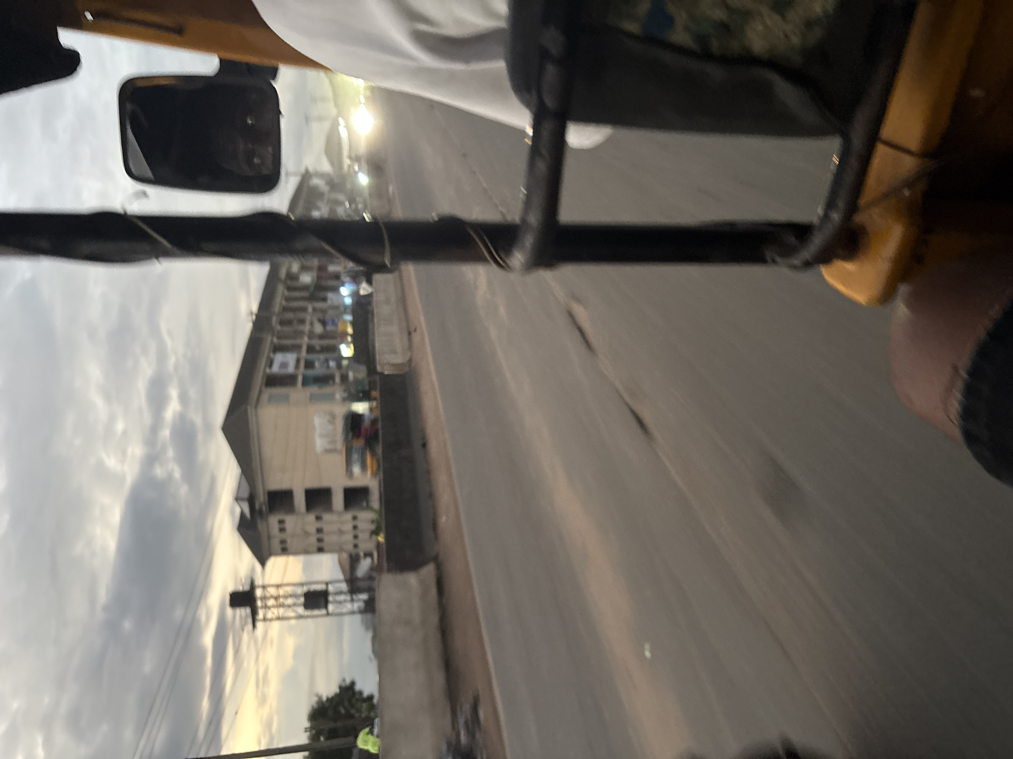 This image captures a street scene in Onitsha, Nigeria, likely during the late afternoon or early evening due to the overcast sky and artificial lights beginning to illuminate. The perspective is from inside a yellow vehicle, possibly a motorized tricycle or tuk-tuk, looking outwards.
In the foreground, the interior of the vehicle is visible, with a dark metal bar from the passenger area and a portion of the yellow exterior. A person's arm and leg are also visible, suggesting the presence of a passenger or the driver.
The mid-ground shows a paved road with lanes. A concrete barrier separates the road from the sidewalk area. Beyond the barrier, a multi-story building with multiple windows is under construction or recently completed. A tall metal structure with water tanks is situated to the left of the building, and a yellow and red vehicle is visible on the road in front of the building. Several streetlights are illuminated, and there are signs of activity along the street, including what appear to be market stalls or shops. The sky is cloudy and gray, with hints of lighter sky near the horizon, indicating the sun is low.