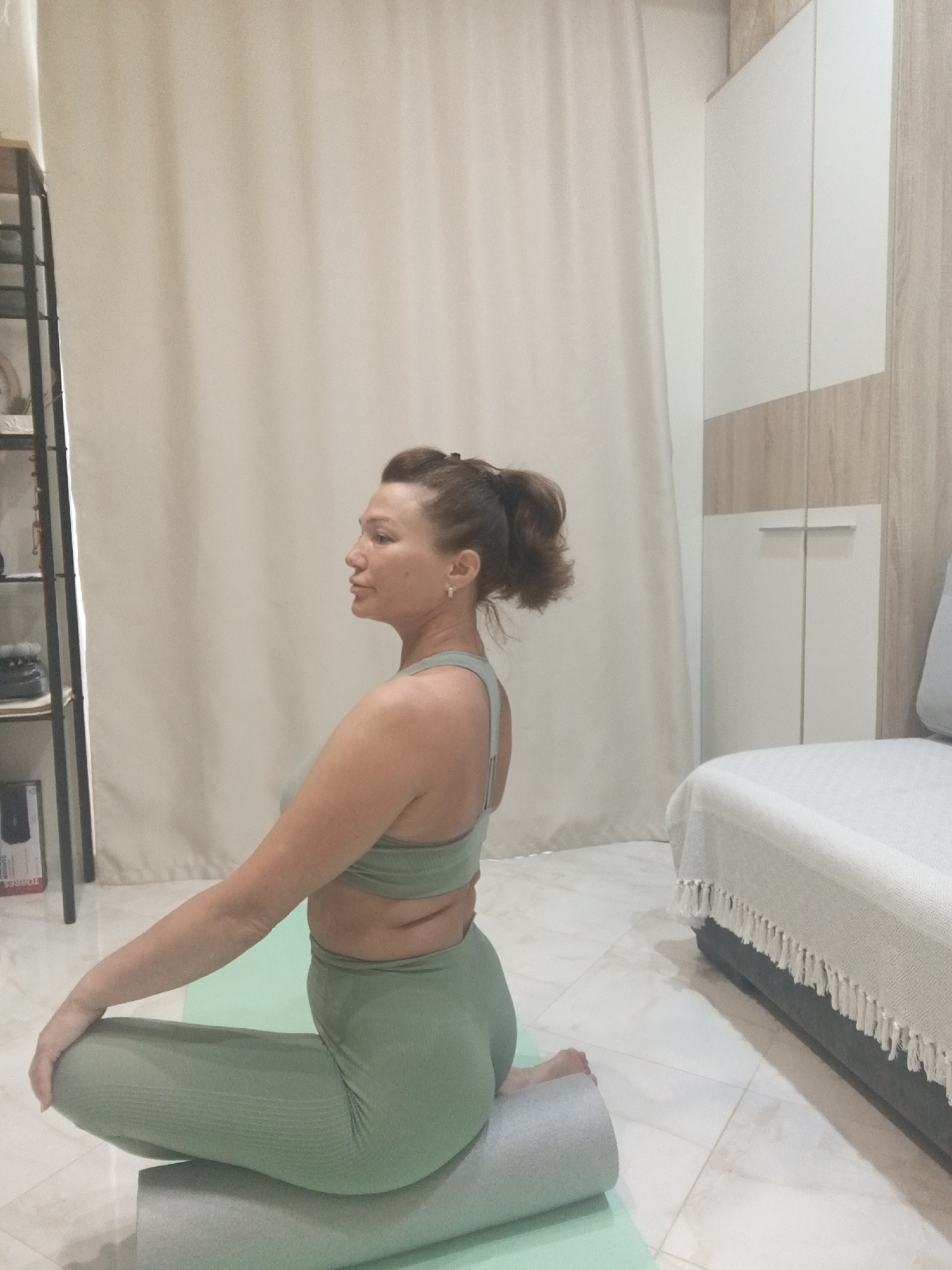 A medium shot captures a Caucasian woman in profile, sitting cross-legged on a yoga mat in a room that appears to be a home. She is wearing a light green athletic top and matching leggings. Her hair is pulled back in a ponytail. The woman is looking to her left, with her right arm resting on her right knee and her left hand on her left knee. A grey foam roller is positioned beneath her. Behind her, to the left, is a black metal shelf unit with various items on it. To the right of the woman is a large white closet with light wood trim. A light-colored couch is visible in the far right of the frame, partially covered by a textured throw blanket with fringe. The floor is tiled with a light, reflective pattern. The overall lighting suggests an indoor setting, possibly during the day. The image does not contain any identifiable landmarks or clues to definitively place it in Sochi, Russia.