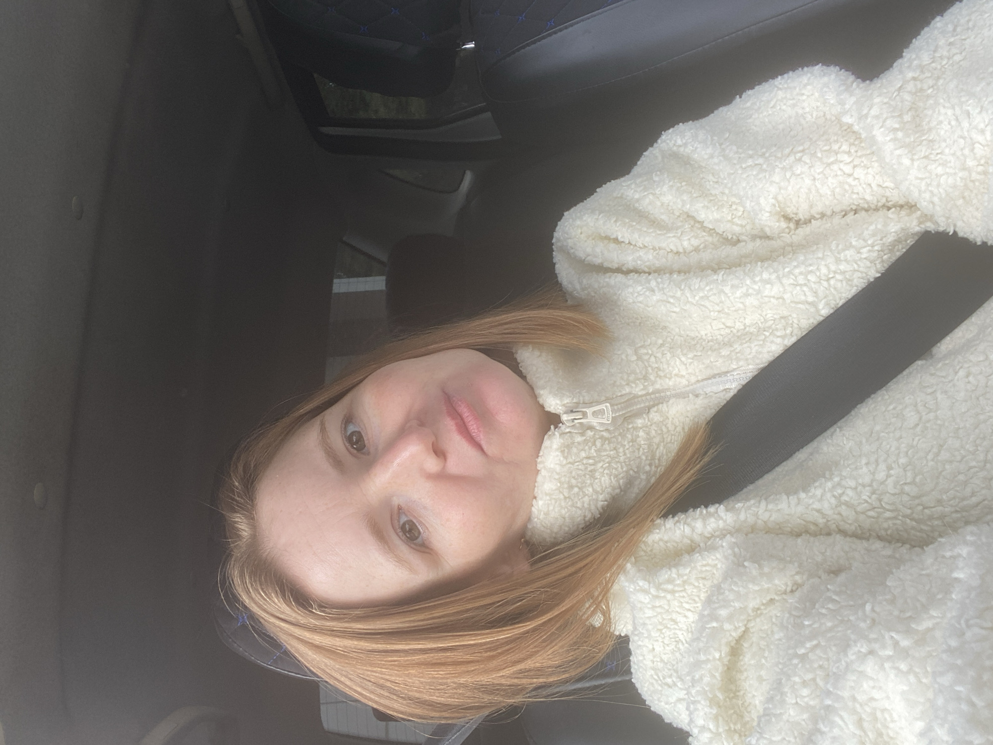 A woman with auburn hair and fair skin is seated in the passenger seat of a car, wearing a cream-colored fuzzy jacket and a black seatbelt. Her head is tilted, and she is looking towards the camera with a neutral expression. The interior of the car is visible, including the dashboard and a dark-colored car seat. The lighting suggests it is daytime, possibly overcast. The image does not contain any identifiable landmarks or contextual cues to determine a specific location within Chaikovsky, Russia.