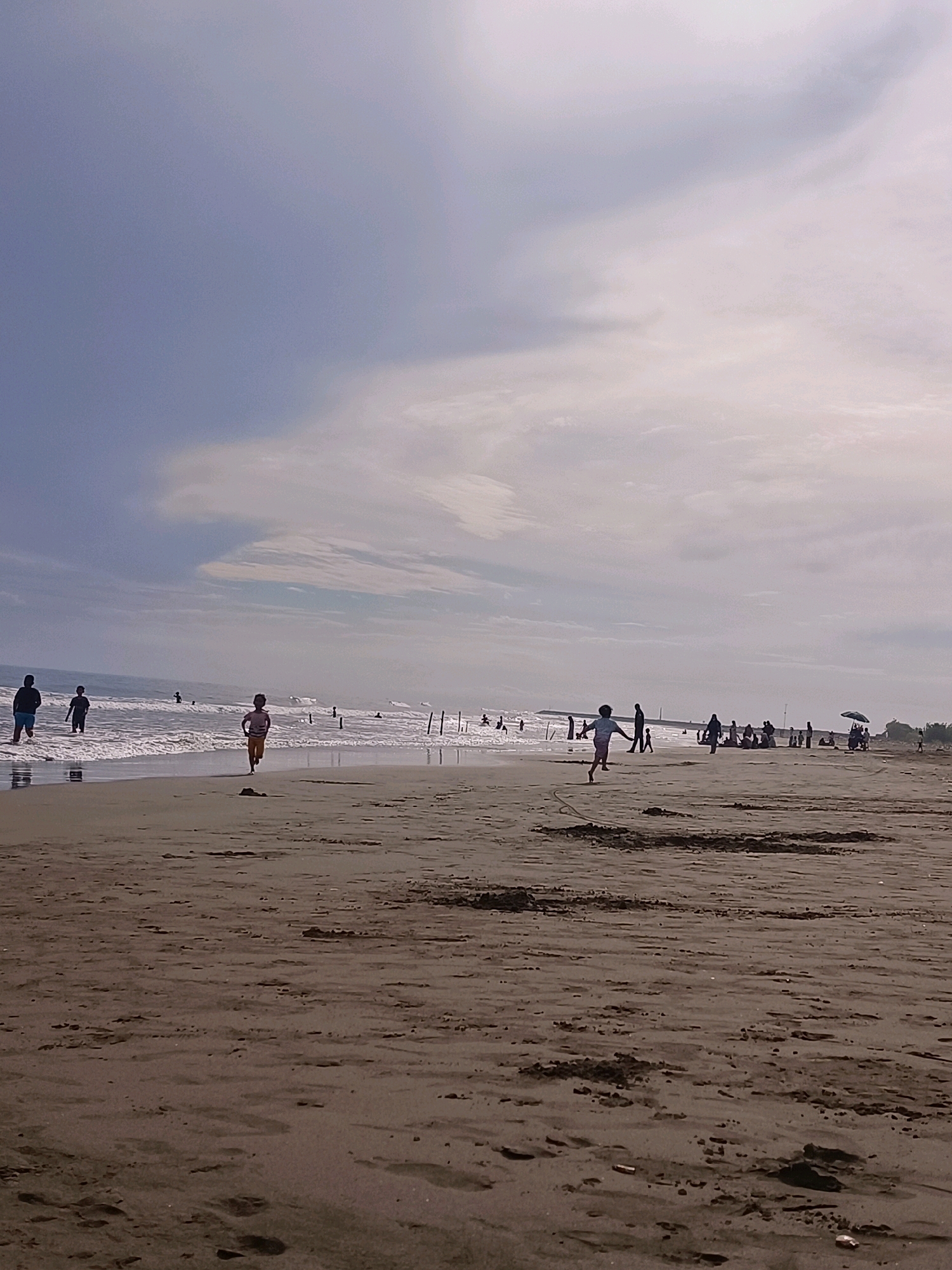 A wide shot captures a beach scene in Reudeuep, Indonesia, under a partly cloudy sky. The foreground is dominated by a sandy beach with scattered debris and footprints. The middle ground features the ocean with gentle waves and several people wading in the shallow water. In the distance, more people are visible along the shoreline, some sitting and others standing. A few vertical poles and an umbrella are also present. The overall impression is of a leisurely day at the beach with various individuals and small groups enjoying the coastal environment.