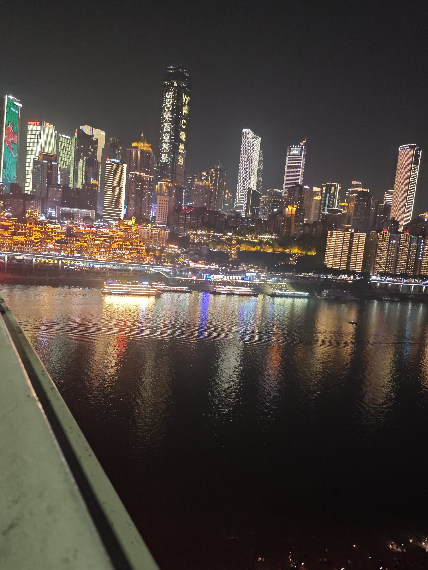 Captured at night, the scene reveals a cityscape reflecting on a dark body of water. High-rise buildings dominate the background, illuminated with bright lights and colors, including some with vertical text displays. The waterfront is lined with glowing structures, with the lights of boats reflecting in the water. A portion of a gray bridge or structure is visible in the lower left corner of the frame. The scene depicts a bustling urban area, potentially in the Yuzhong District of China, as suggested by the image's context.