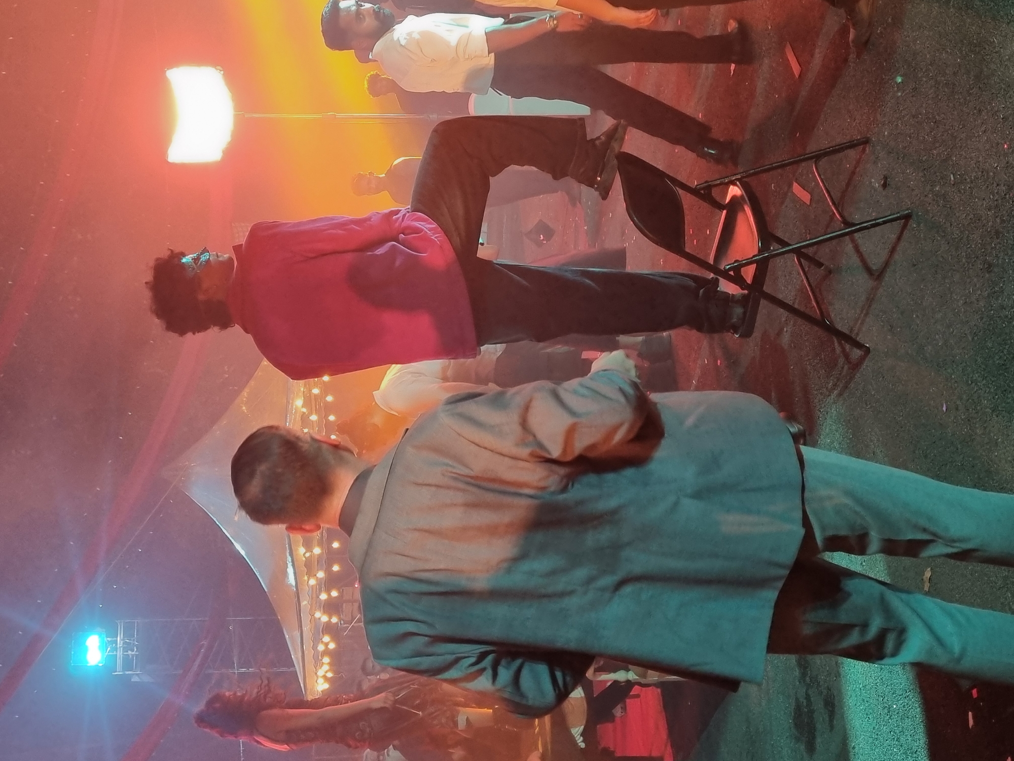 The image depicts an indoor or covered night event, likely a film set, photoshoot, or stage performance, located in Kalamassery, India. The scene is dominated by artificial lighting, creating a dramatic atmosphere.
Several individuals are present. In the foreground, a man in a grey suit stands with his back to the viewer. To his left and slightly behind, another man with dark, curly hair and glasses wears a vibrant pink/magenta shirt and dark pants, appearing to look towards a bright light. Further back, another man in a white shirt and dark trousers also faces the direction of the light. A fourth person, possibly seated, has their legs visible, suggesting they are also part of the arrangement.
A prominent bright rectangular white light on a stand illuminates the central figures, casting a warm glow. In the background, a blue stage light is visible, and warm string lights or fairy lights are draped across a translucent, tent-like structure, adding to the event's ambiance. A black folding chair lies on its side on the dark, textured ground, which appears to be concrete or pavement, with some red confetti-like debris scattered across it. The collective arrangement of people and lighting suggests a carefully composed visual production. No visible text is present in the image.