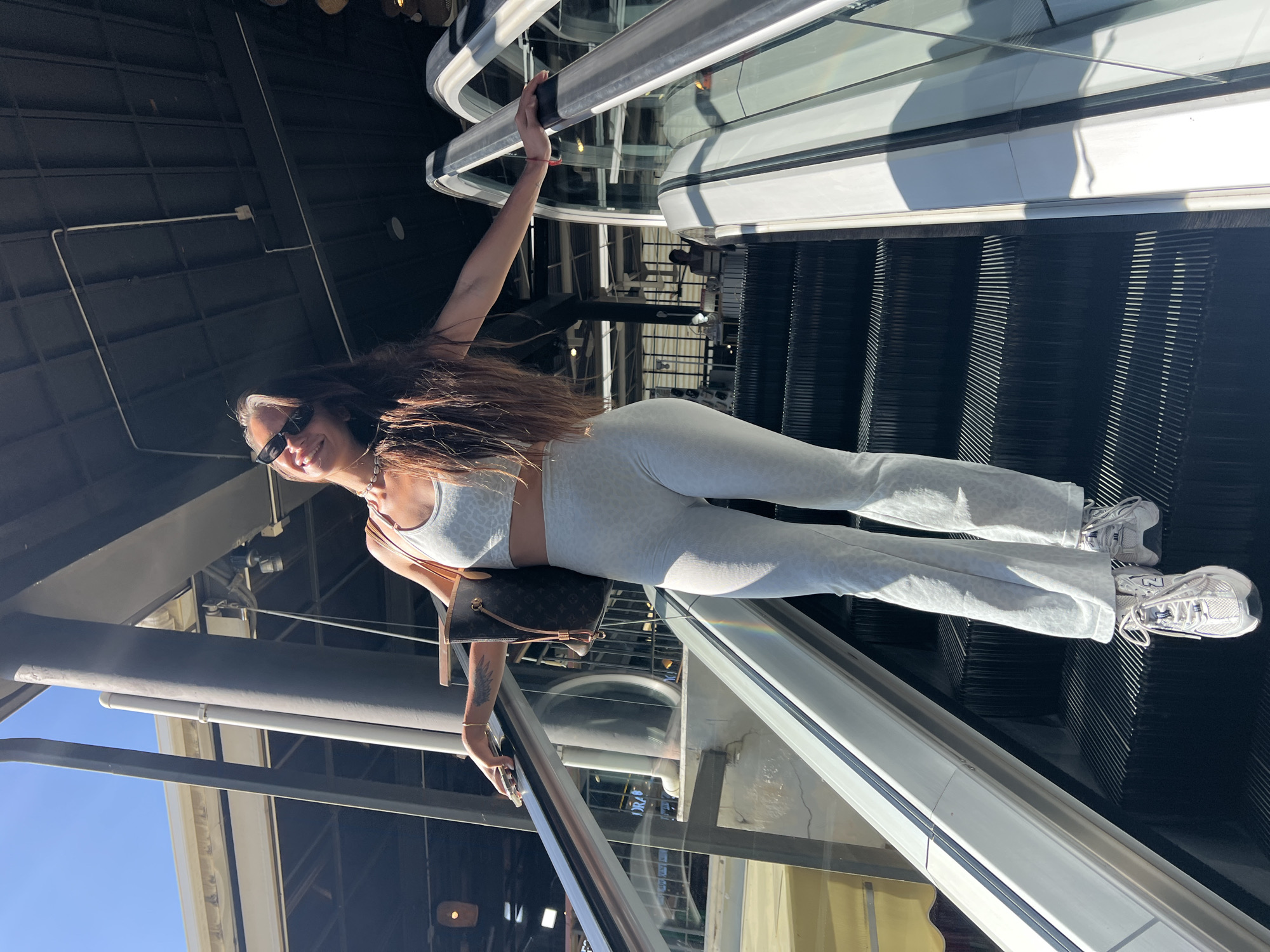 A woman stands on an escalator at an indoor location in บ้านบางรักษ์, Thailand. She wears a light-colored athletic-style outfit, sunglasses, and white sneakers. She also has a purse. She is holding onto the escalator railing with both hands. The escalator is moving upward. The location has a modern architectural style with exposed metal beams and glass.
