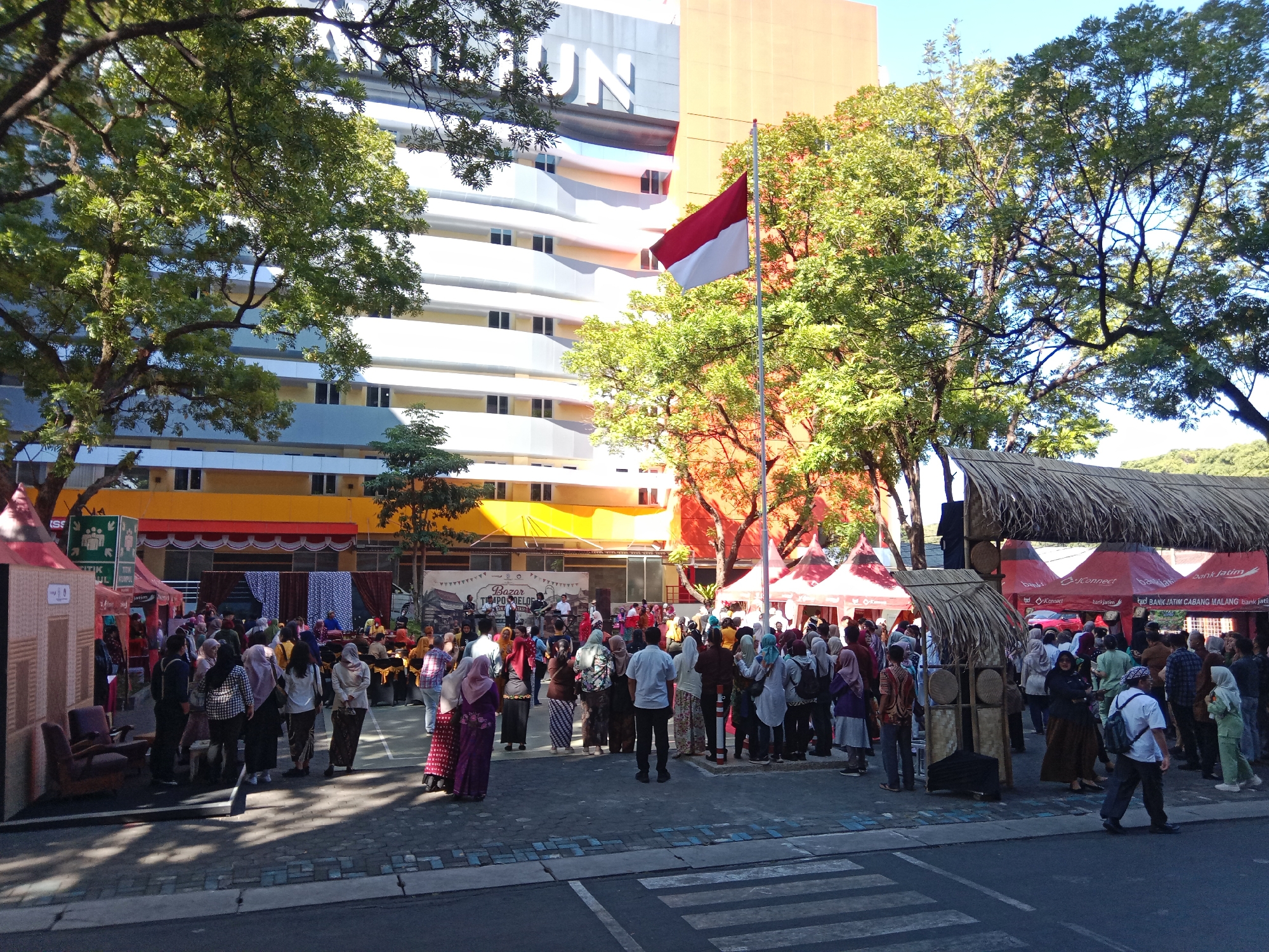 This is a wide shot of an outdoor event in front of a multi-story building. The building has white and yellow horizontal bands and appears to be an institutional building, possibly a university, with the letters "UN" visible near the top. An Indonesian flag flies on a pole in front of the building, to the right of a large tree with green foliage.
In the foreground, a street with a crosswalk is visible. The event itself features several temporary stalls and tents, many with red canopies and some with straw roofs. A crowd of people, dressed in a mix of casual and traditional attire, are milling about, browsing the stalls, and standing in groups. Some individuals are walking, while others appear to be talking and socializing. There are several chairs and a small, makeshift structure resembling a house to the left.
In the background, behind the stalls, there's a stage with a backdrop that has a pattern of blue and white rhombuses and a banner that reads "BAZAR TEMPO MELO". Some people are gathered around the stage, where a band appears to be performing. The overall atmosphere suggests a lively community event or market. The presence of the "bank Jatim" logo on some tents indicates a local bank sponsor. The location is likely a public space in the city of Malang, Indonesia, given the bank's name and the cultural context of the event.