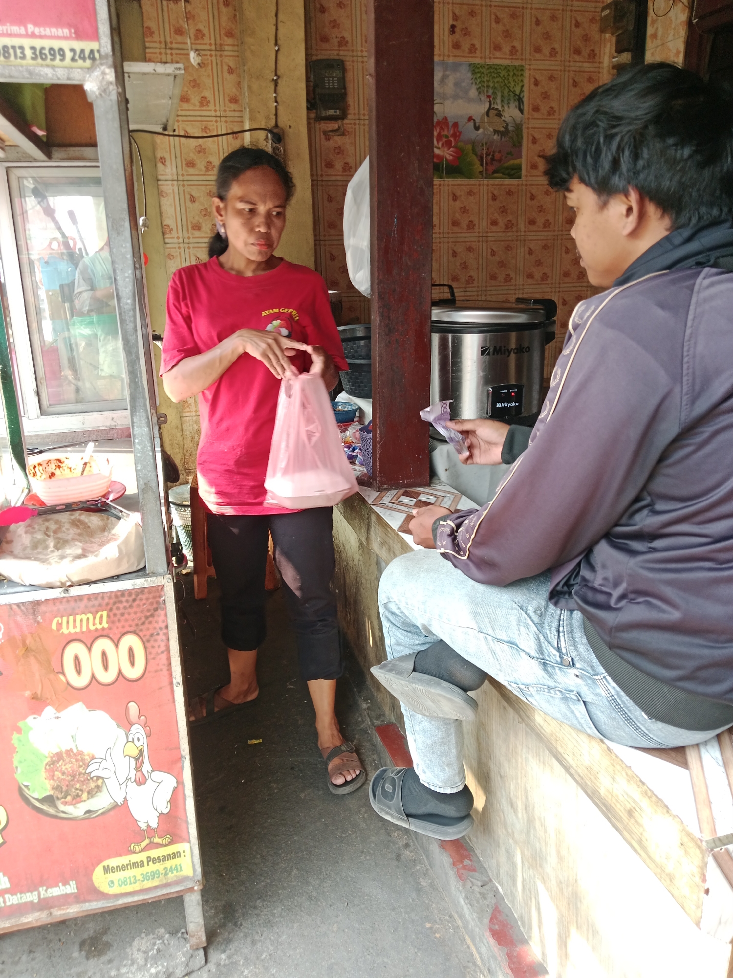 A woman in a red t-shirt and black pants stands on a dirt floor in front of a food stall. She is holding a pink plastic bag in her right hand. A man in a dark jacket and light blue jeans sits on a wooden bench to her right, holding a bill in his left hand. Behind them, a rice cooker with the brand name "Miyako" is visible. The stall has a red sign with a cartoon chicken and text indicating "cuma 000". The wall behind the stall is covered in patterned tiles. The scene appears to be in Kediri City, Indonesia.