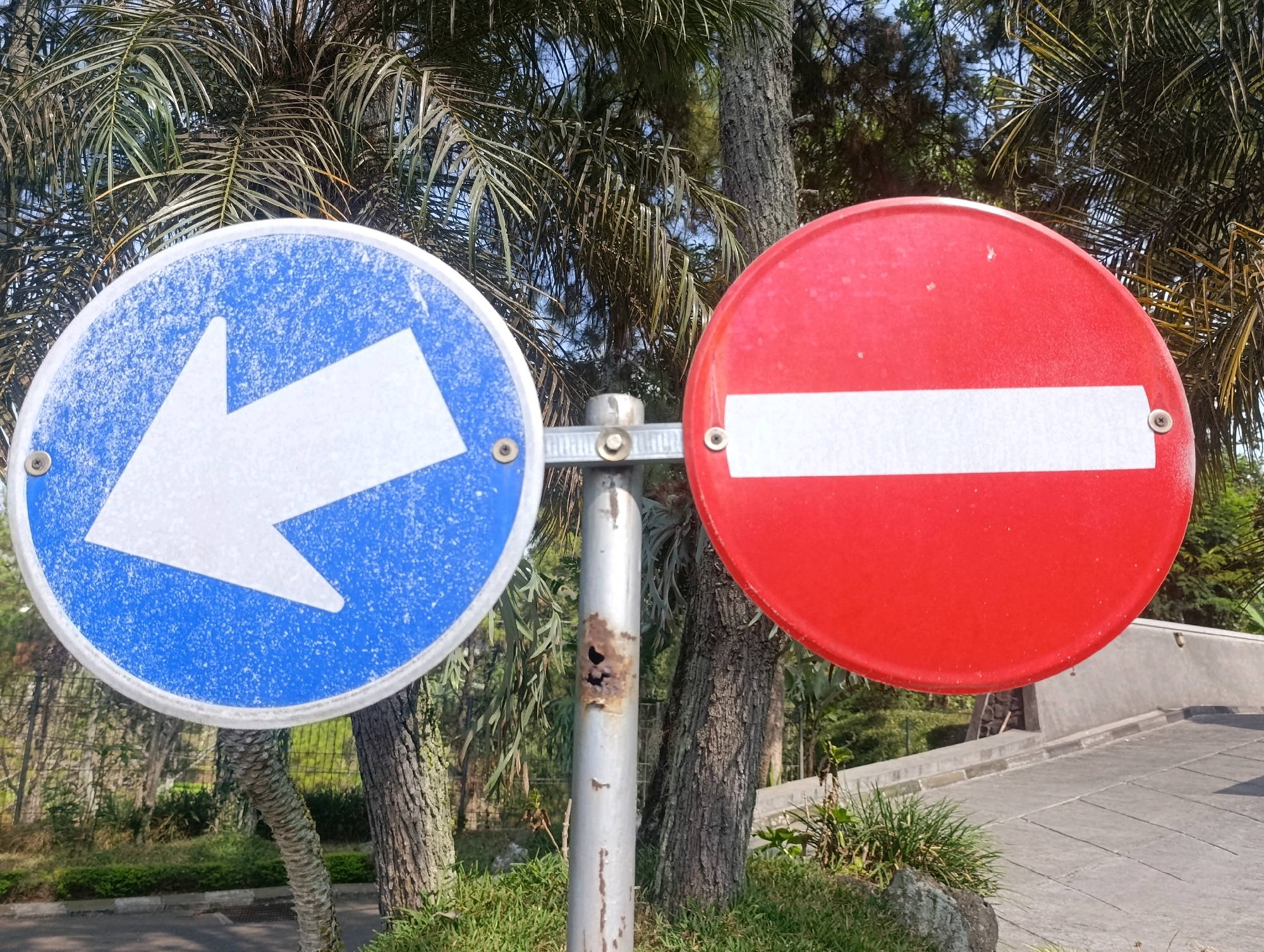 The image shows two circular traffic signs mounted on a metal pole in Cihanjuang, Indonesia. The sign on the left is blue with a white arrow pointing to the left. The sign on the right is red with a white horizontal bar. Both signs appear weathered. The pole supporting the signs is silver and shows signs of rust and damage. In the background, there are palm trees, other trees, and greenery, suggesting an outdoor, possibly park-like or roadside environment. A paved pathway is visible on the right side of the image. The lighting suggests it is daytime.