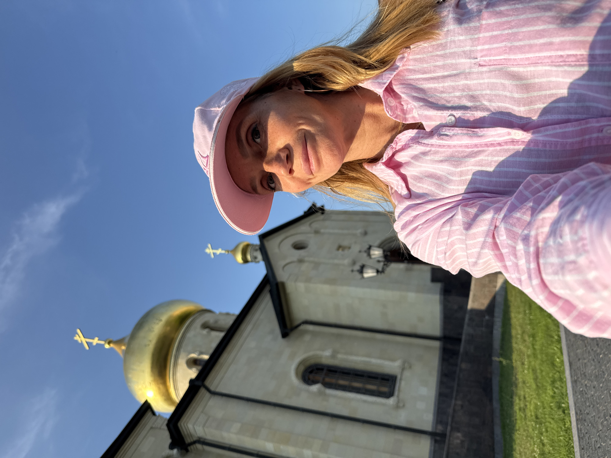 A medium shot captures a woman with blonde hair wearing a pink baseball cap and a pink and white striped button-down shirt, standing outdoors with a church and a clear blue sky in the background. The woman is smiling and looking slightly off-camera to her right. The church, with its light-colored stone facade and a prominent golden dome topped with a cross, is visible behind her on the left side of the frame. The ground in front of the church appears to be paved with cobblestones and a strip of green grass. The image was taken in Kirishi, Russia.