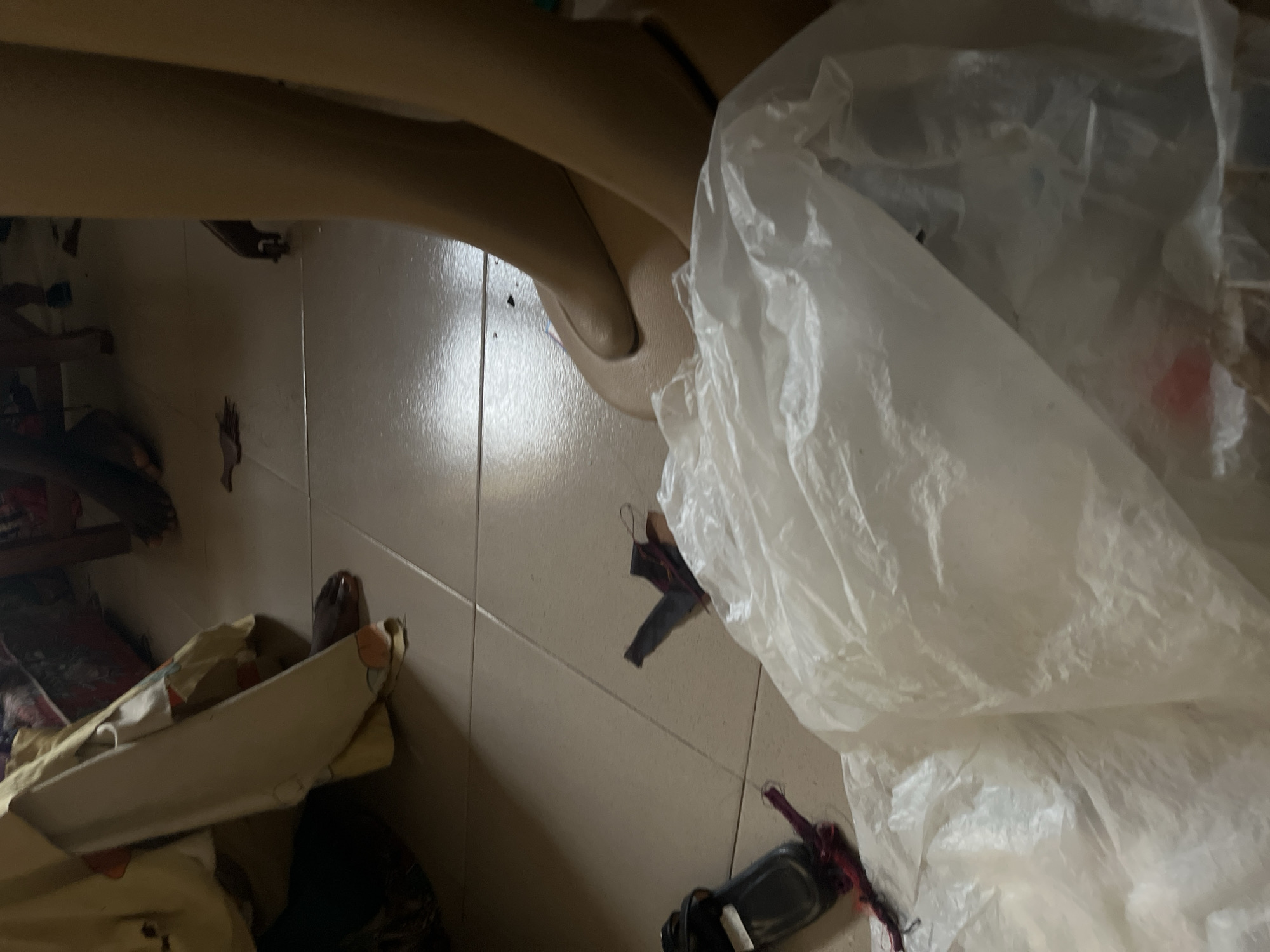 The image depicts a cluttered floor in what appears to be an indoor setting, possibly a room in Jauro Yinu, Nigeria. The floor is tiled with large, light-colored squares. Scattered across the floor are various items, including a black sandal with red threads attached, a crumpled white plastic bag or sheeting, and what looks like a dark, flat object, possibly a piece of fabric or debris. In the background, parts of furniture and other objects are visible, as well as several feet and legs. Some of the feet are bare, while others appear to be wearing shoes or are partially obscured. The lighting suggests it may be evening or a dimly lit interior. The overall impression is one of disarray and untidiness.