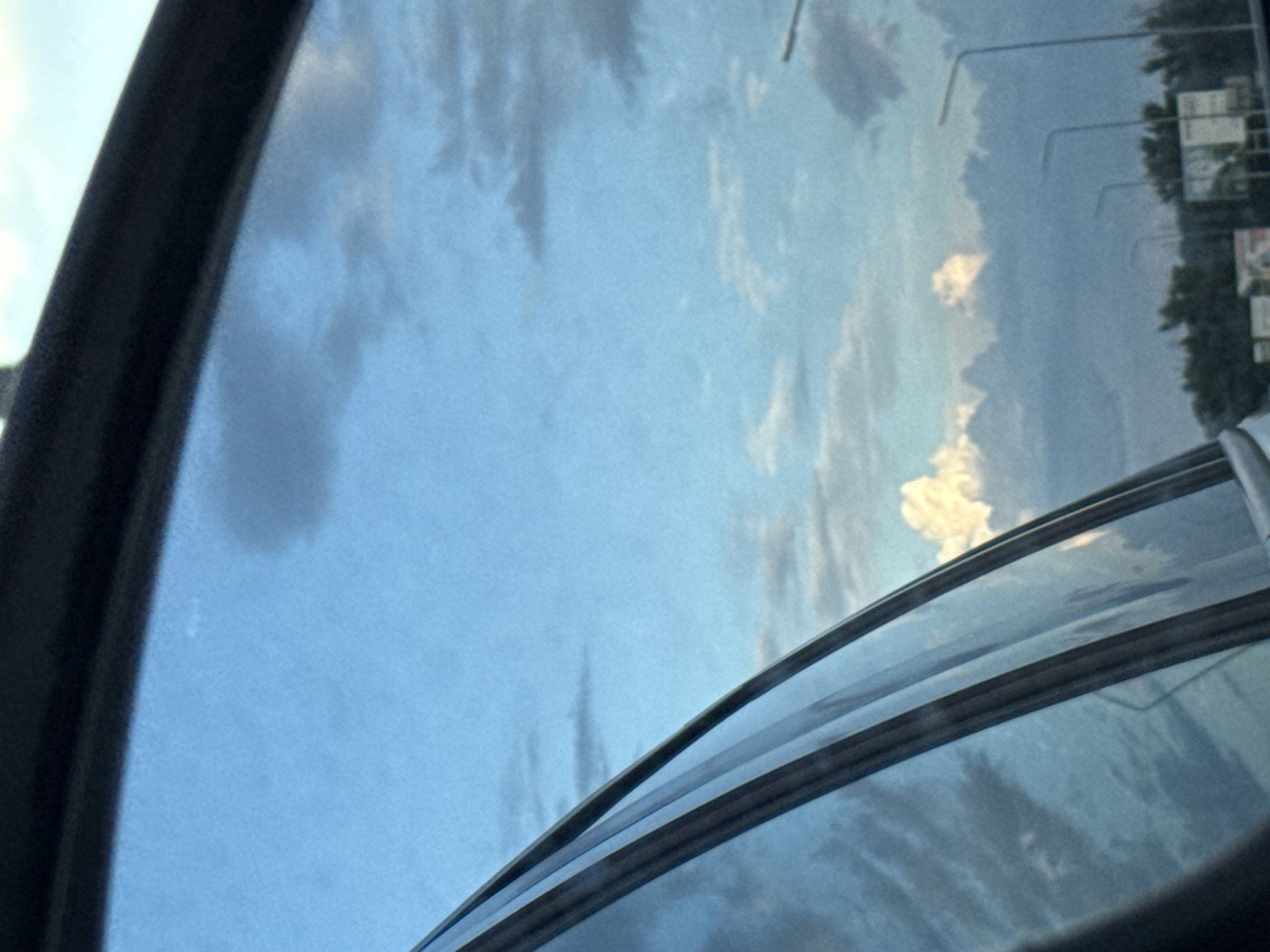 The media file shows a view through what appears to be a car window or sunroof. The primary subject is the sky, with a mix of blue sky and white, fluffy clouds. The clouds are scattered and appear to be in motion, suggesting wind. In the reflection on the glass, several light poles and trees are visible, indicating an outdoor setting, likely a roadside environment. There are also some rectangular signs visible in the reflection, though their text is illegible. The overall lighting suggests it is daytime, possibly late afternoon or early morning given the soft light and the presence of clouds. The scene conveys a sense of travel or observation from a stationary or moving vehicle. There are no people visible. The setting appears to be a somewhat rural or suburban area based on the presence of trees and light poles. The image is taken from a low angle, looking upwards towards the sky. The colors are dominated by blues and whites, with hints of green and darker tones from the reflections.