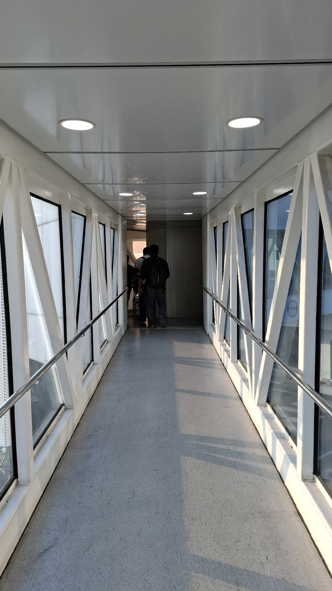 The image shows a person walking down a covered jet bridge at an airport. The jet bridge has a white ceiling with circular lights, white triangular supports on the sides, and large windows. The floor is gray. The person is wearing a dark shirt, gray pants, and a backpack. They are walking away from the camera towards a closed door at the end of the jet bridge. The lighting suggests it is daytime, with sunlight casting shadows on the floor. The scene is sterile and functional, typical of an airport environment.