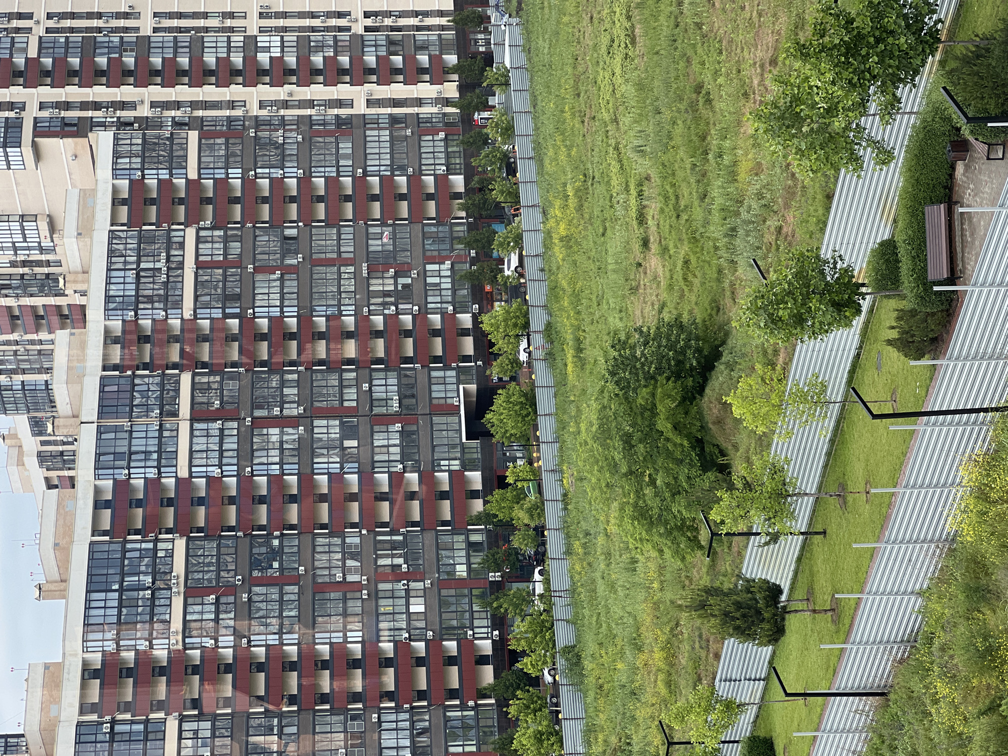 The image shows a high-angle view of a park area in Krasnodar, Russia, juxtaposed with a large apartment building. On the left side, the apartment building is tall with a repetitive pattern of windows, balconies, and decorative elements. The building appears modern, with many units having air conditioning units.
To the right of the building, there is a green park area. A pathway runs through the park, featuring benches, trees, and modern light fixtures. The grass is thick and green, and there appear to be several walkways with seating areas. A fence separates the park from the road. Cars are parked along the edge of the road.