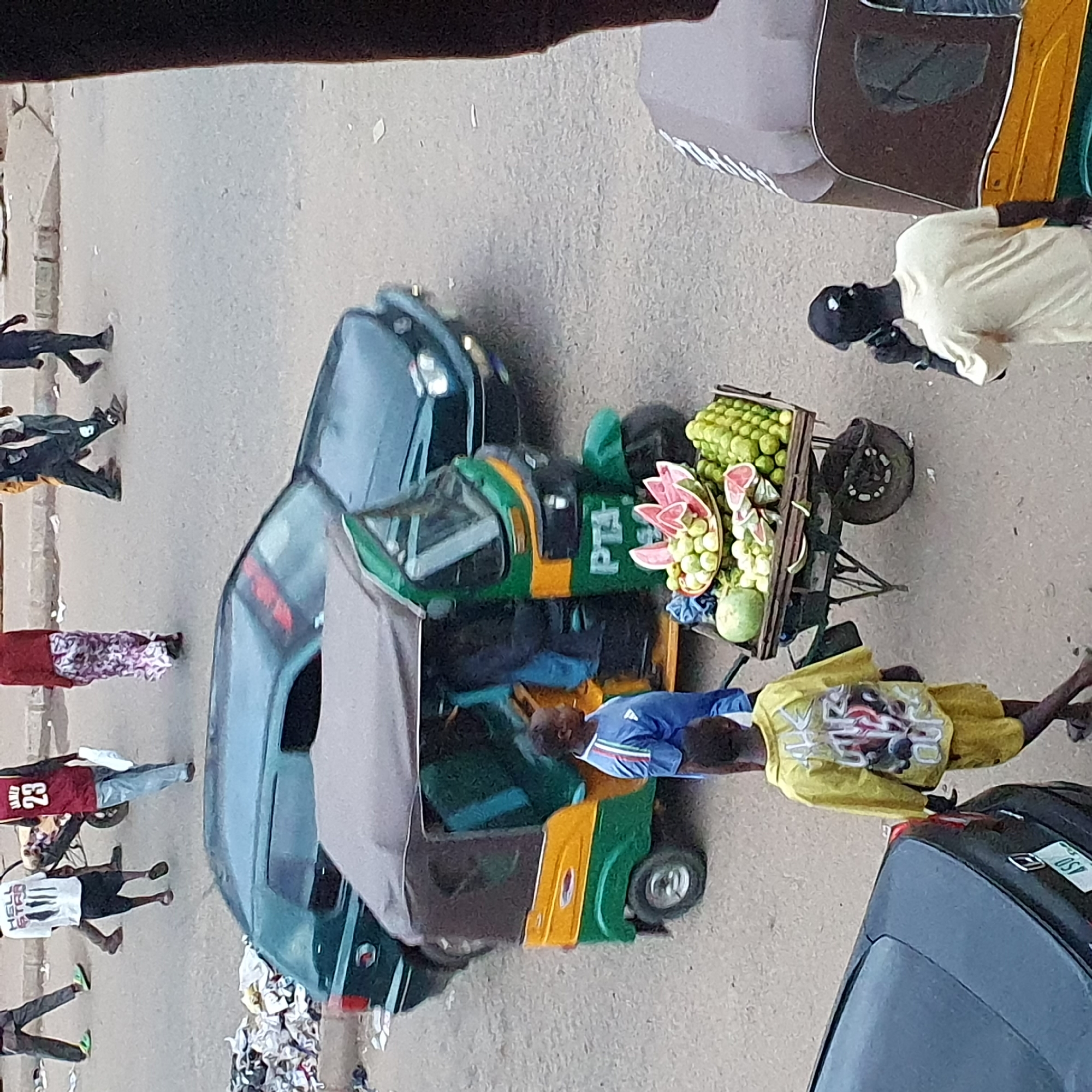 In an outdoor urban environment, possibly a market or street, the scene captures a snapshot of daily life. A three-wheeled auto-rickshaw, painted in green and yellow, is parked on a paved surface, a common mode of transportation and mobile vendor in many parts of the world. In front of the auto-rickshaw, a cart is laden with a variety of fruits, including watermelons cut into slices and a generous amount of green citrus fruits, likely limes or small oranges, and other green fruits.
Several people are present in the image. A young boy, wearing a bright yellow jacket and shorts, stands to the left, his back to the camera. Another person, wearing a blue t-shirt with white and red stripes, is partially visible near the auto-rickshaw. Further in the background, other individuals are seen walking, some with shopping bags. One person is on the phone, while others are dressed in casual attire. The presence of other vehicles, including a dark-colored car and a larger vehicle partially visible on the right, suggests a busy street setting. Debris, including what appears to be discarded clothing and paper, is scattered on the ground, indicating a less-than-pristine urban environment. The lighting suggests it is daytime.