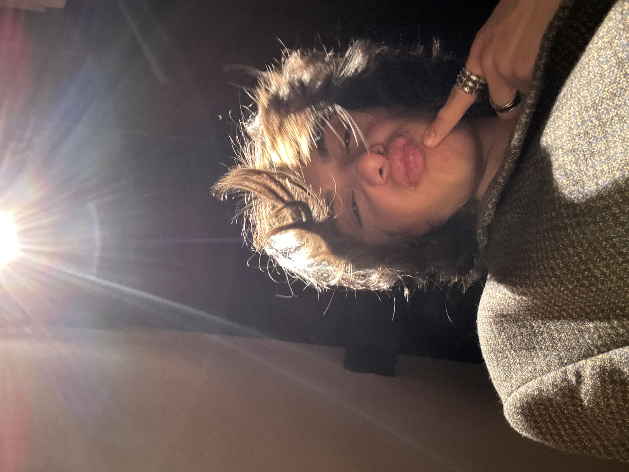 The image shows a person with brown hair and a grey tweed jacket posing for a selfie. The person is making a kissing face and has a finger on their cheek, adorned with several rings. The background is dark, with a bright light source causing a glare. There is no information to suggest this image was taken in Yerevan, Armenia.