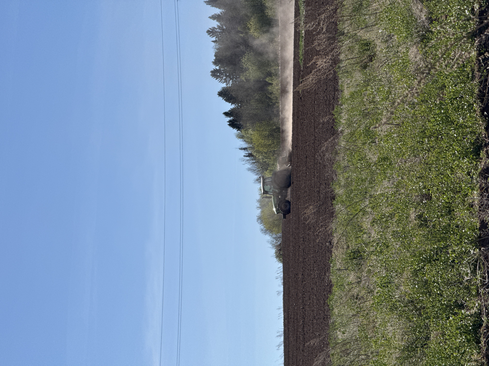 A tractor is plowing a field in Лобаново, Russia, creating a dust cloud behind it. A forest borders one side of the field, and a grassy area the other. Power lines are visible against the clear blue sky. The field is plowed with dark brown soil.