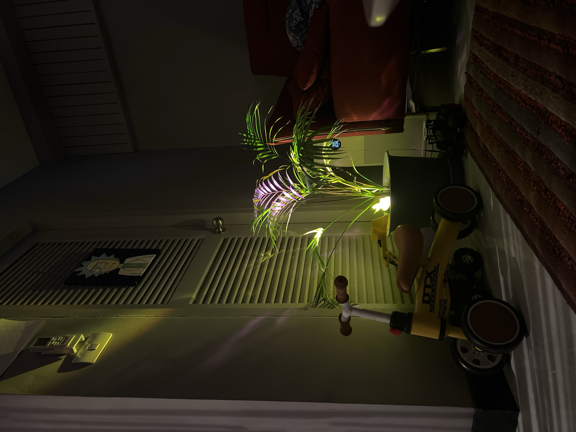 The image, taken indoors at night or in the evening, shows a domestic scene illuminated by artificial light. A prominent potted plant, likely a palm, stands in the center, its fronds glowing with green and purple light. In the foreground, a yellow children's ride-on toy, possibly a balance bike, is visible, featuring brown accents and the text "DTX GEAR BIKE" on its side.
To the left, a louvered door is affixed with a black plaque depicting a cartoon character resembling Rick from "Rick and Morty." Below the door, on a shelf or surface, two white remote controls are placed. In the background, a red armchair or sofa is partially visible. Near the plant, an electronic unit, possibly an air conditioner or humidifier, displays digital numbers "24" and either "70" or "76." A patterned rug with stripes in shades of brown and orange occupies the right side of the frame. The scene is static, with no people or activity present, conveying a quiet, domestic atmosphere.