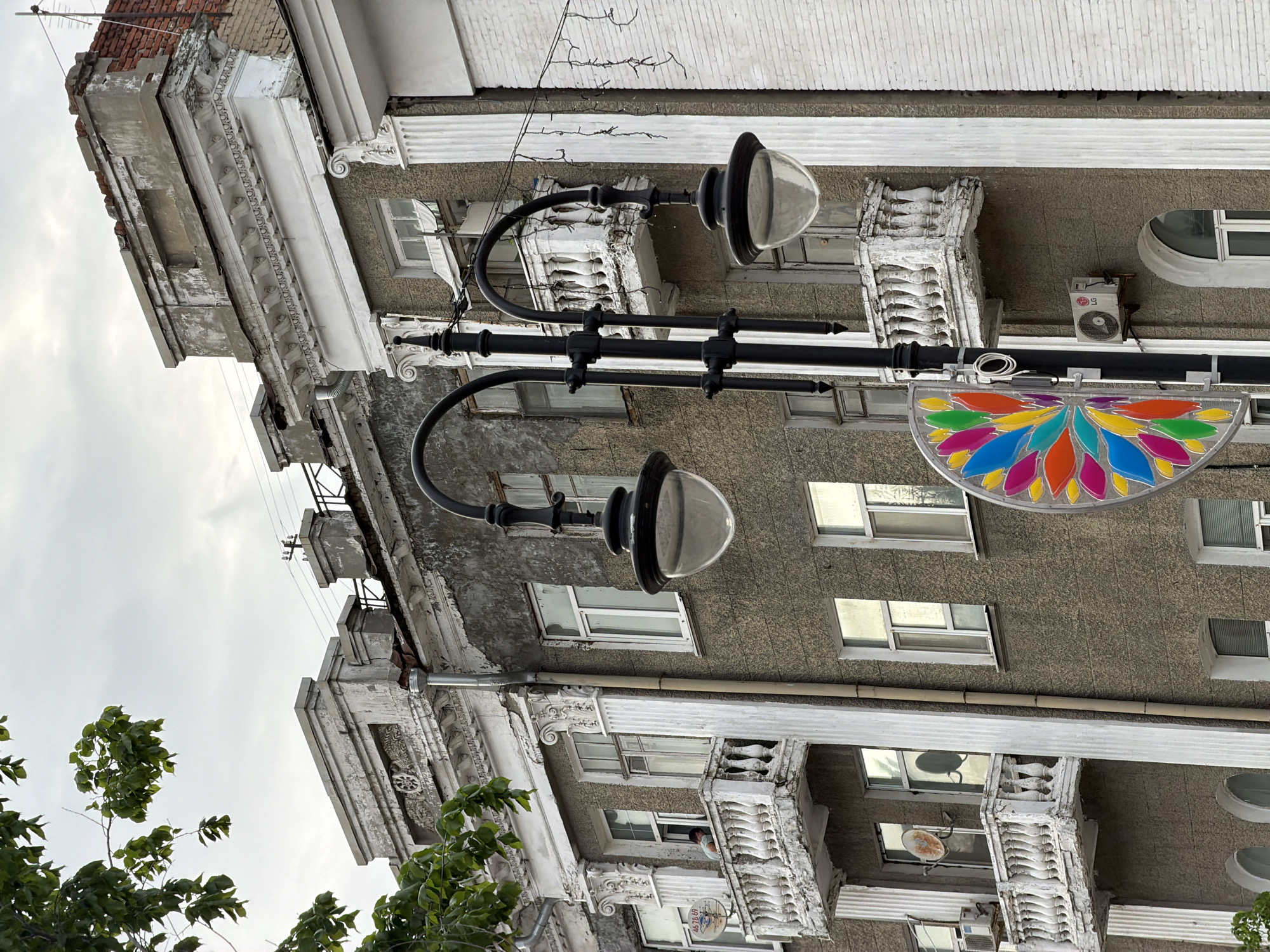 The image shows a multi-story building in Saratov, Russia. The building's exterior is characterized by weathered grey stucco, white decorative architectural elements, and windows of various sizes. A black, double-lamp street light is mounted on the building facade, with a colorful stained-glass sign hanging nearby. The sky is overcast. A tree is visible on the bottom left of the frame.