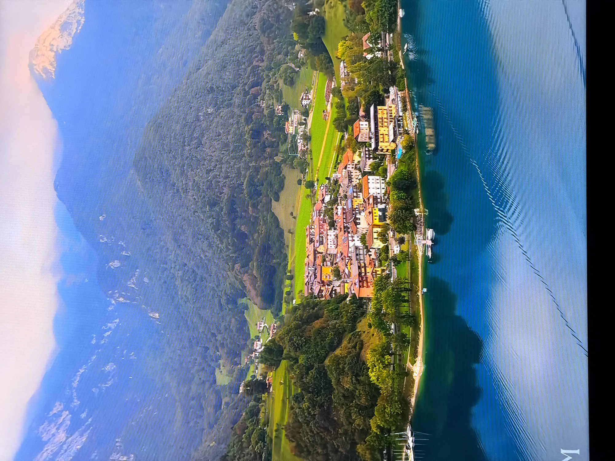 This is an aerial drone shot of a scenic landscape. In the foreground, a vast expanse of deep blue water with subtle ripples reflects the sky and surrounding environment. Along the shoreline, a small town is nestled against a backdrop of lush green hills and majestic mountains. The town features buildings with terracotta roofs, characteristic of European architecture. The hills are covered with dense forests and interspersed with green meadows. In the distance, towering mountains rise, with their peaks shrouded in soft, white clouds, and one peak catches the warm glow of sunlight. The water in the foreground is calm, with the reflection of the sky creating a soft, ethereal effect. There are a few boats docked by the shore, suggesting recreational activities. The overall scene evokes a sense of tranquility and natural beauty. There is no information or indication that this image is located in Chirchik, Uzbekistan.