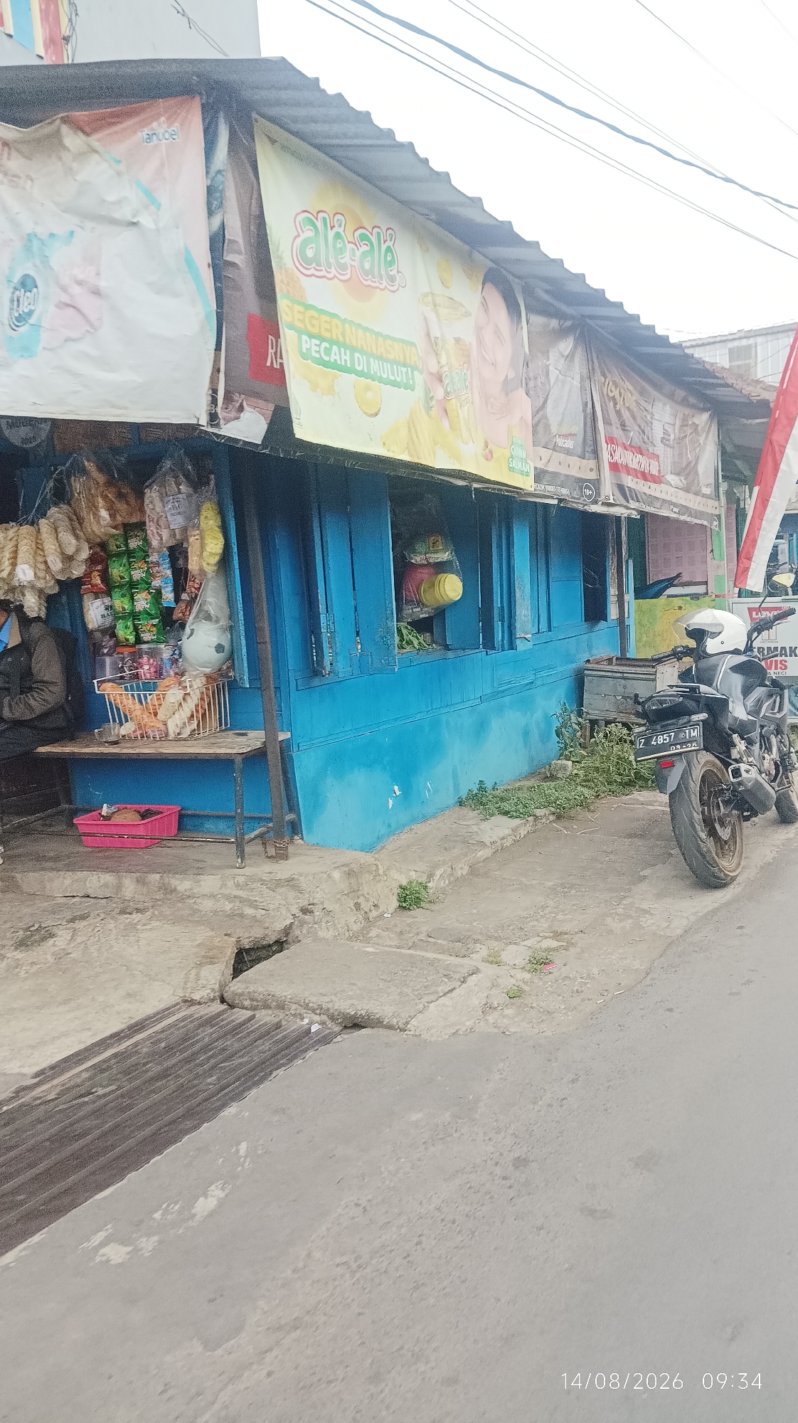 The image depicts a street-side stall in Tasikmalaya, Indonesia, with a weathered blue wooden structure serving as the storefront. A man in a dark jacket is seated to the left, observing the scene. The stall displays various packaged goods, including snacks and drinks, hanging from the ceiling and arranged on shelves. A basket filled with goods sits on a small wooden table in front of the stall. A pink plastic container is on the ground beneath the table. A motorcycle with a white helmet is parked on the right side of the road, in front of the stall. Banners advertising "Ale-ale" juice and other products are displayed above the stall. The ground in front of the stall is paved with concrete and shows signs of wear and tear, with a drain visible in the lower left corner. The street is asphalted. The timestamp 14/08/2026 09:34 is visible in the bottom right corner of the image.
