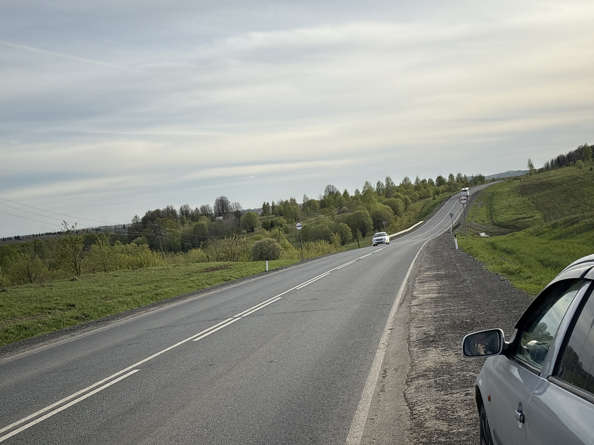 The image depicts a rural road scene near Бикбарда, Russia. The road is paved and has double solid white lines indicating no passing. Green fields and trees line both sides of the road, with some small hills in the background. The sky is overcast with various shades of gray and white clouds. There are at least two cars visible on the road, one closer and one further away. A roadside sign with a white circle and a horizontal line indicating a prohibition is present. A silver car is partially visible on the right side of the frame, with its side mirror showing a reflection. The ground immediately adjacent to the road appears to be a mix of gravel and dirt.