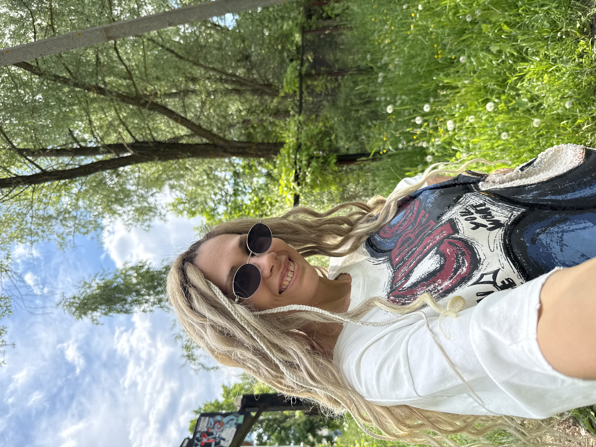 A selfie captures a smiling woman outdoors, appearing to be in Заволжье, Russia. She looks directly at the camera with a broad, joyful smile. Her long, blonde hair is styled in numerous dreadlocks or thick braids, some incorporating lighter strands. She wears dark, round-framed sunglasses.
She is dressed in a white short-sleeved t-shirt featuring a prominent, abstract graphic design in red, blue, and black. A dark-colored jacket or hoodie, possibly black or dark blue with a white, fluffy interior lining, is draped over her shoulders. On the jacket's design, some stylized text is visible, including what appears to be "YOU WANT" and other fragmented words.
The background depicts a lush natural environment under a bright, sunny sky. Tall trees with vibrant green foliage occupy the upper and left parts of the frame, suggesting a park or forest. The ground is covered in rich green grass, dotted with numerous white dandelion seed heads and some small yellow flowers. The sky above is blue with scattered white clouds, indicating clear weather. In the lower-left corner, a low fence or structure, adorned with graffiti, is partially visible. The overall mood of the image is cheerful and natural, capturing a moment of outdoor enjoyment during daytime.