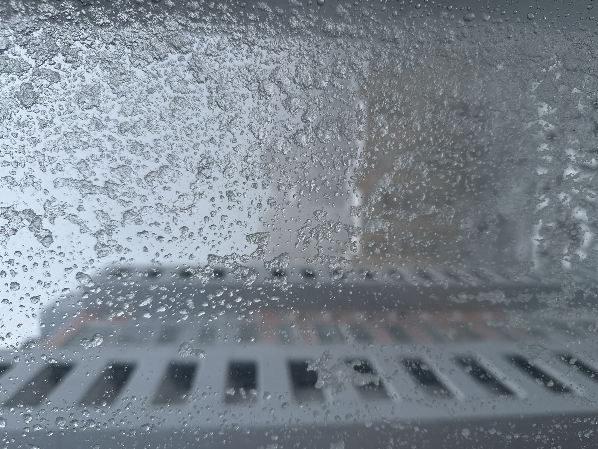 The image shows a view through a window covered in ice and water droplets. A metal grid-like structure is visible in the lower portion of the frame. Through the frosted window, a building can be partially seen in the background. The overall scene suggests a cold and possibly snowy environment. The location, Moscow, Russia, is only inferred and cannot be definitively confirmed based on the visual information alone.