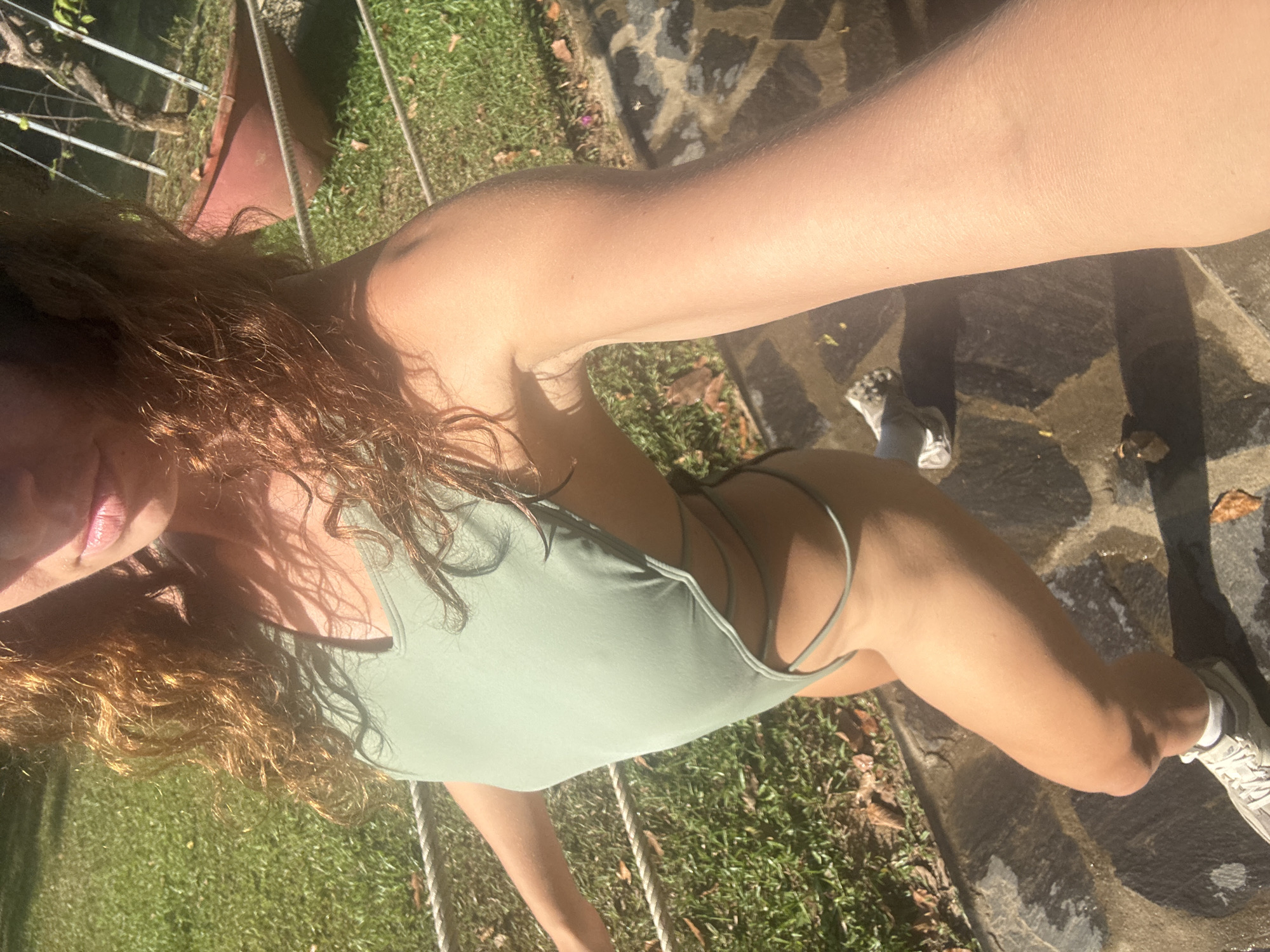 A person is taking a selfie. The subject is a light-skinned individual with curly auburn hair, wearing a light green one-piece swimsuit. The person's left arm is extended toward the camera. The setting is outdoors, with grass and a rock-covered ground visible. Ropes are running on the ground as well. A shoe is seen on a foot in the background. The location of the photo is unspecified, so the city of Đà Nẵng, Vietnam, cannot be confirmed.