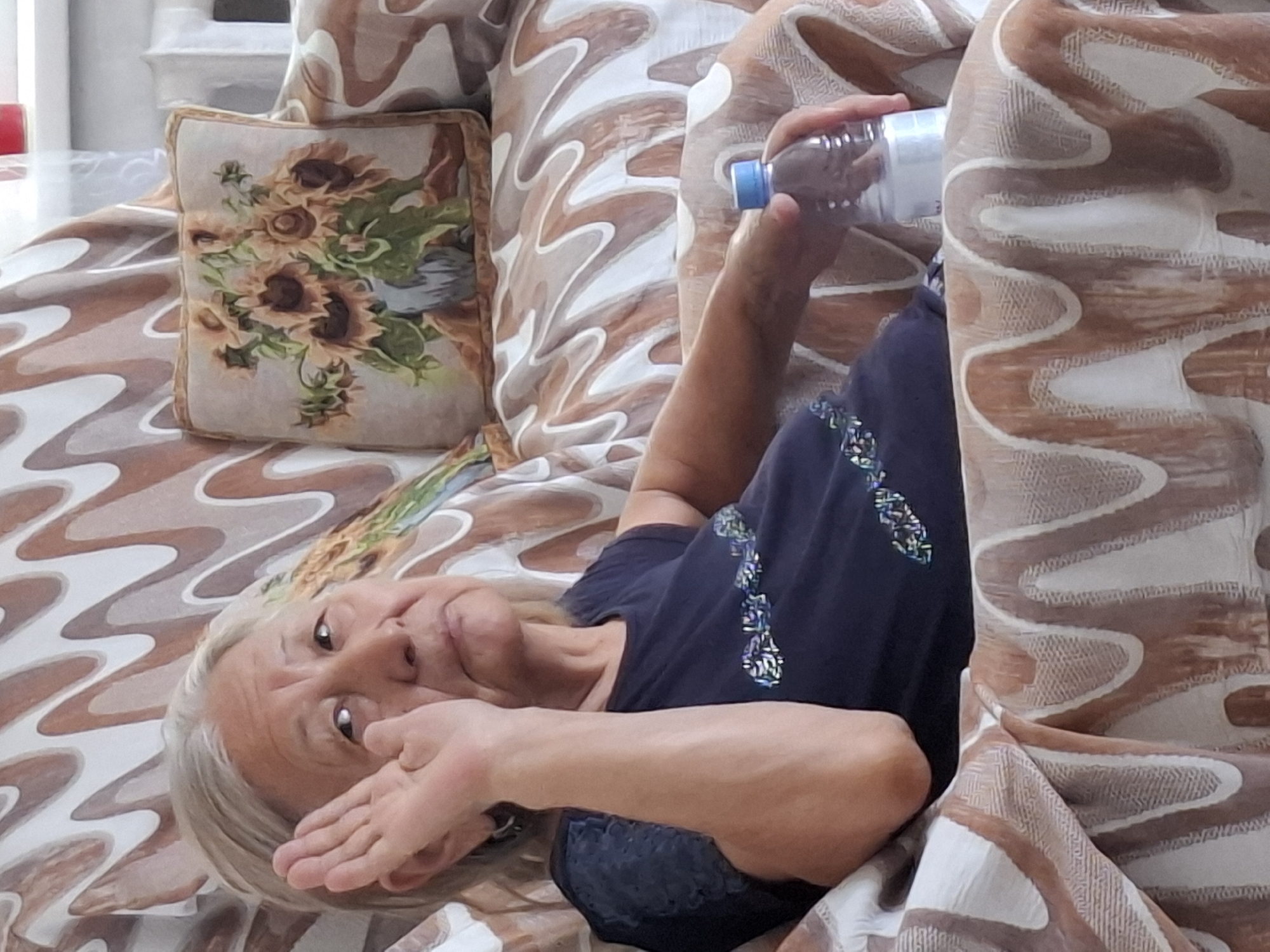 An elderly East Asian woman with gray hair and fair skin is sitting on a couch in what appears to be a residential interior. She is wearing a dark blue t-shirt with a decorative embellishment across the chest. Her left hand is raised to her face, with her palm open and facing outward. Her gaze is directed slightly upward and to her left, with a neutral expression. Next to her right arm, on the couch, is a clear plastic water bottle with a blue cap. The couch is covered in a fabric with a swirling pattern of brown and off-white wavy lines. Behind her, on the couch, is a decorative throw pillow featuring an illustration of sunflowers in a vase. The background is blurred but suggests an indoor setting with soft lighting. The image does not contain any specific landmarks or details that would allow for precise identification of its location within Almaty, Kazakhstan.