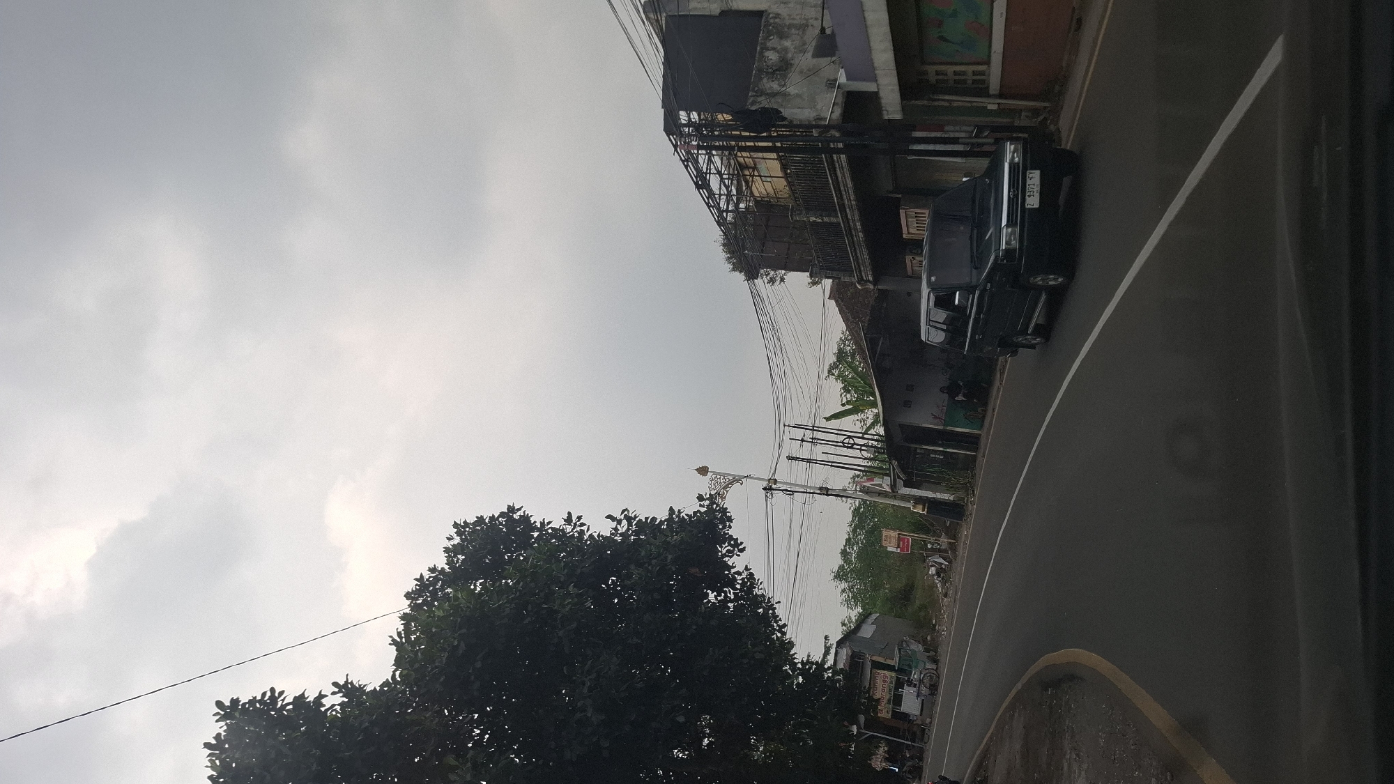 The image shows a street scene in the city of Cilaku, Indonesia, under an overcast sky. A dark-colored van is traveling on the road, which curves to the left. On the right side of the road, there are several buildings, some appearing to be shops or residences, with visible electrical wires and poles. A large tree with dark green foliage is on the left side of the road, partially obscuring the view of some smaller buildings and a bicycle parked on the side. The road has white lane markings. The overall impression is that of a typical street in a populated area of Indonesia.