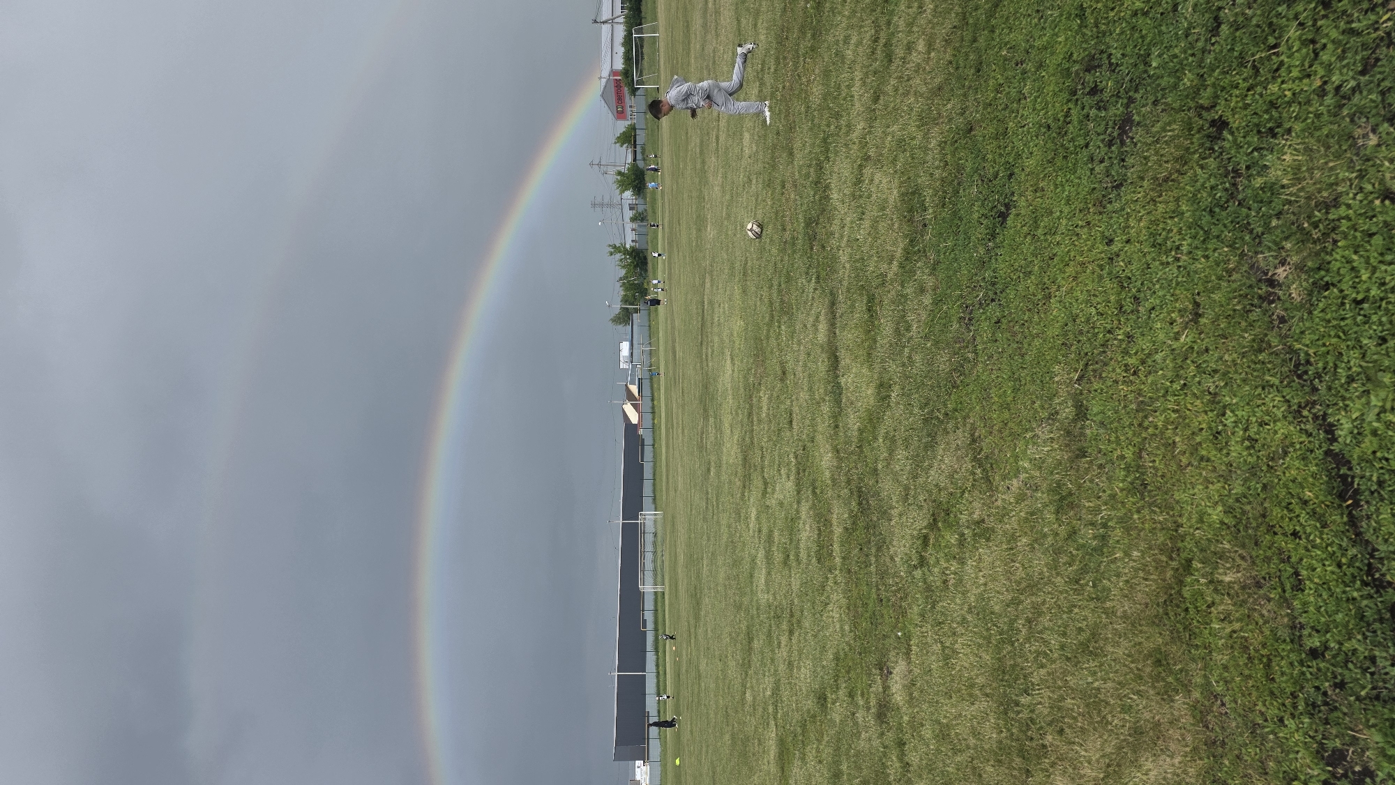 An outdoor sports field is depicted under a sky displaying a double rainbow. The image is oriented vertically, but the content suggests a landscape view. A prominent rainbow, vibrant with colors, arches across the left side of the frame, extending from a dark, cloudy sky towards the grassy field. A fainter, secondary rainbow is visible above it.
On the bright green grass of the field, a person wearing light grey athletic attire and white shoes is captured in a dynamic, mid-action pose, possibly diving or falling, with their body nearly parallel to the ground. A soccer ball rests on the grass nearby, slightly to their left. In the background, other smaller figures are sparsely distributed across the expansive field. White goalposts stand at either end of the field, and in the far background, there are several buildings, including one with a distinctive red roof, along with trees and utility poles with power lines. The field itself shows variations in grass texture and color, with some areas appearing more lush than others. Faint, unidentifiable text can be seen on a building in the background. The presence of the rainbow strongly suggests recent rainfall while the sun is still out, likely indicating late afternoon or early evening.
