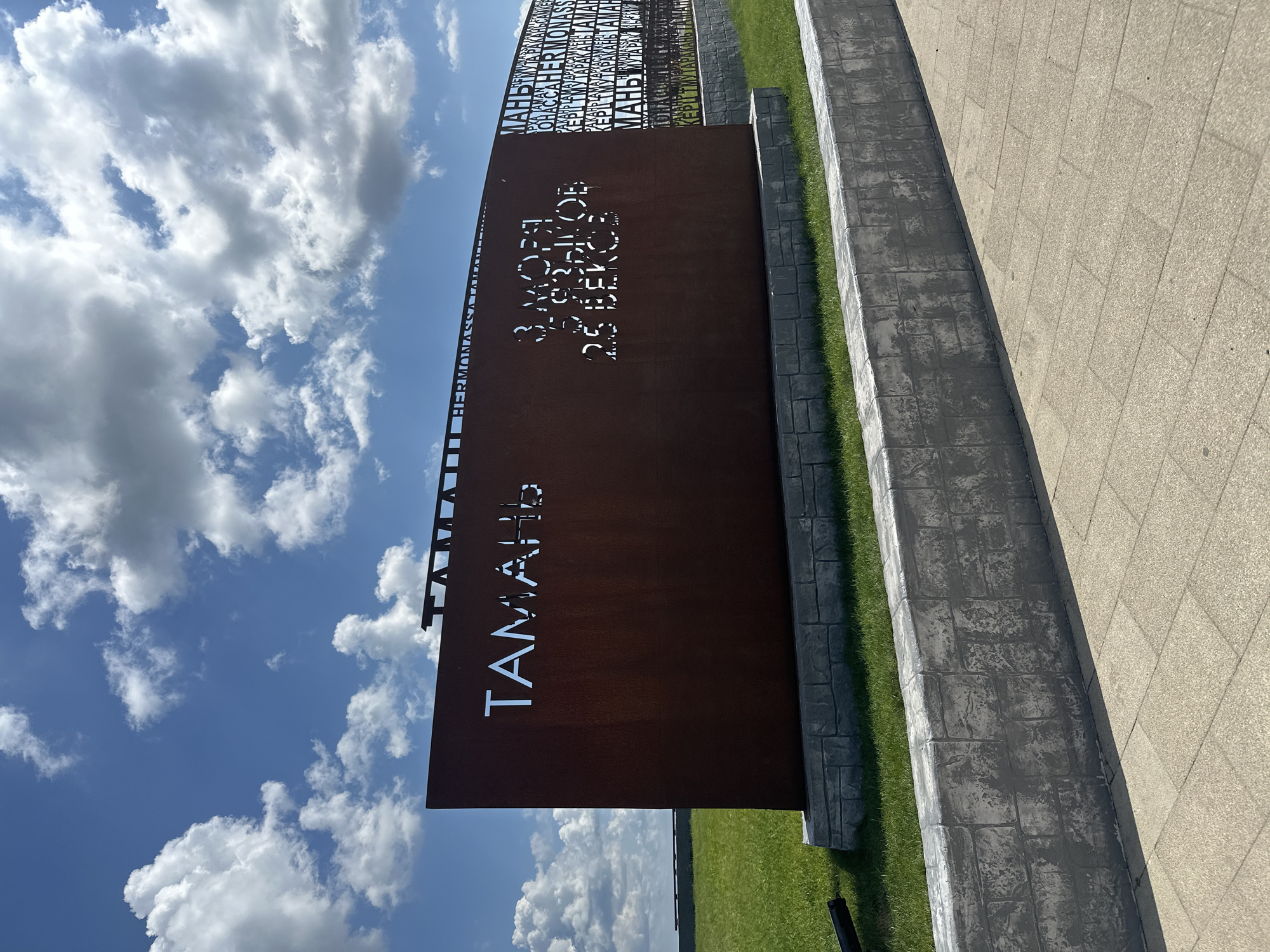 The image captures an outdoor monument in Taman, Russia, under a bright blue sky with scattered white clouds. The monument is a large, rust-colored metal structure with cut-out lettering. The most prominent text reads "ТАМАНЬ" in large, white, capital letters on the left side of the structure. To the right of this, smaller text is also cut out, including "3 МОЯ 309 КОРОВ 25 ВЕКОВ".
The monument is supported by a base that mimics stone blocks, surrounded by a manicured green lawn. A grey, textured concrete wall runs in the foreground, separating the lawn from a paved walkway. In the background, a fence-like structure with repeating text, also cut out from metal, is visible. The overall scene suggests a historical or cultural landmark. There are no people visible in the image.