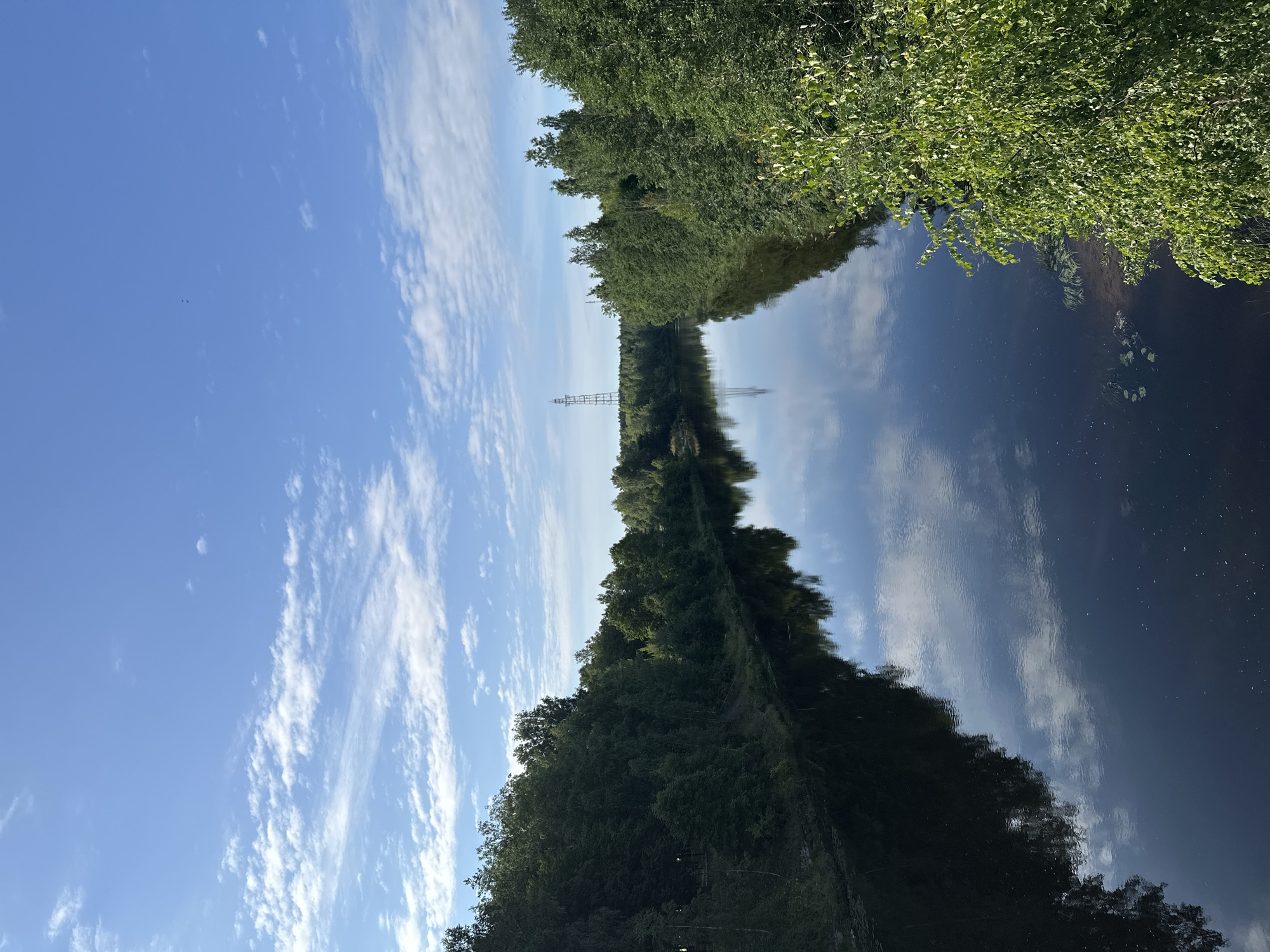 An aerial view captures a calm river flowing through a densely forested area under a clear blue sky with scattered white clouds. The river's dark surface perfectly reflects the trees lining its banks and the sky above, creating a mirror image. In the distance, a tall electrical pylon stands amidst the trees. The foliage is lush and green, suggesting a temperate climate, possibly during the summer or early autumn. The scene evokes a sense of tranquility and natural beauty. Based on the provided information, this landscape likely represents the natural surroundings of Slantsy, Russia, a city situated along the Luga River.