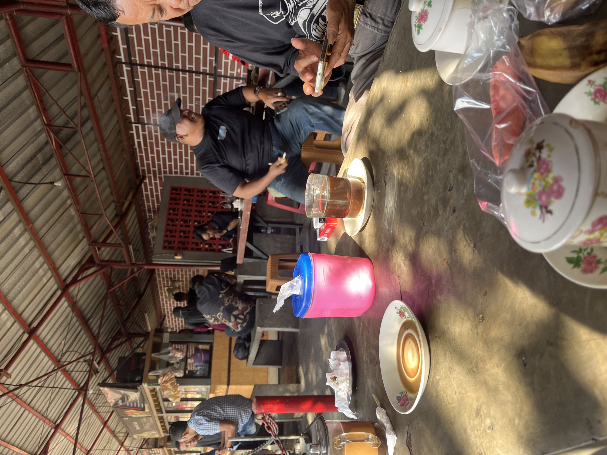 In a covered outdoor seating area, likely a roadside eatery in Singosari, Indonesia, several men are gathered. A man in the foreground, on the right, is engrossed in his smartphone, while a man next to him, wearing a black t-shirt and a baseball cap, smiles and holds a cigarette. In the middle ground, a man in a checkered shirt sits at a table. A pink tissue box and a glass of iced tea are prominent on a textured table in the foreground. The background reveals a brick wall, a counter area with packaged goods, and other patrons seated at tables. The scene is illuminated by natural sunlight, casting distinct shadows.