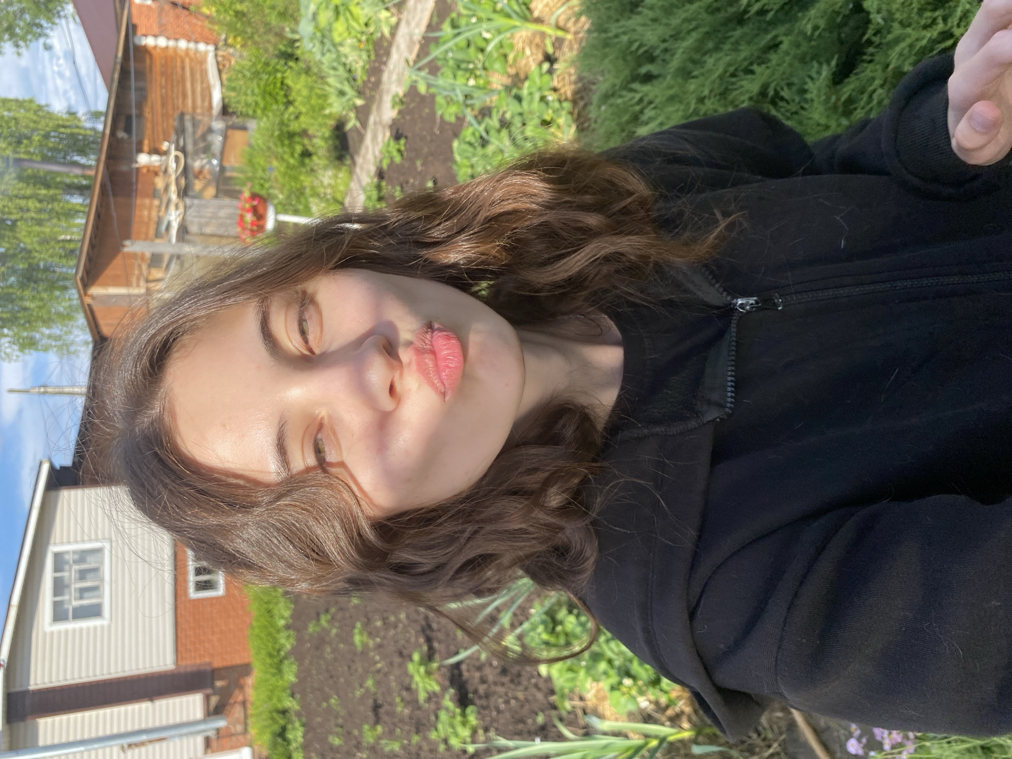 A young woman with brown hair and light skin wears a black zip-up hoodie and pouts her lips for a selfie. The background shows a garden with green plants and dark soil. Wooden houses with red roofs and white siding are visible in the distance. The location is likely Votkinsk, Russia based on user information.