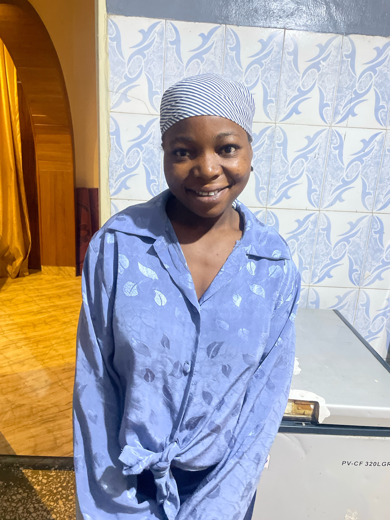 A young woman is standing and smiling at the camera. She is wearing a blue and white striped headscarf and a light blue button-up shirt with a leaf pattern. The shirt is tied in a knot at her waist. She is standing in front of a tiled wall with a blue and white pattern. To her left, there is a yellow curtain and an arched doorway. To her right, there is a portion of a white refrigerator with text that reads "PV-CF 320LGR". The lighting in the image is warm and appears to be artificial. The location is identified as the city of Jos, Nigeria.