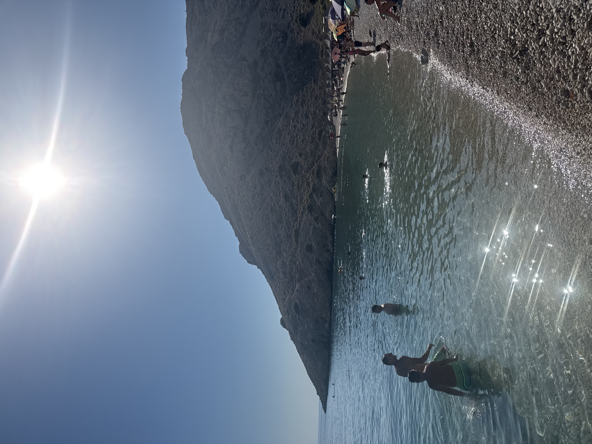 The image depicts a sun-drenched beach scene in Stara Baška, Croatia. A large, rocky hill dominates the background, casting a shadow over parts of the beach and water. The sun is high in the clear blue sky, creating bright lens flares across the water's surface.

In the foreground, several people are visible in the shallow, clear water. Two men are interacting, one appearing to be talking while standing chest-deep in the water. Another person, possibly a child, is nearby. Further out in the water, other individuals are swimming and enjoying the sea.

The beach itself is composed of smooth pebbles. Along the shoreline, a line of beach umbrellas provides shade, and several people are seated or lying on the beach. The water is calm and reflects the bright sunlight, creating shimmering patterns. The overall atmosphere is one of a relaxed summer day at the coast.