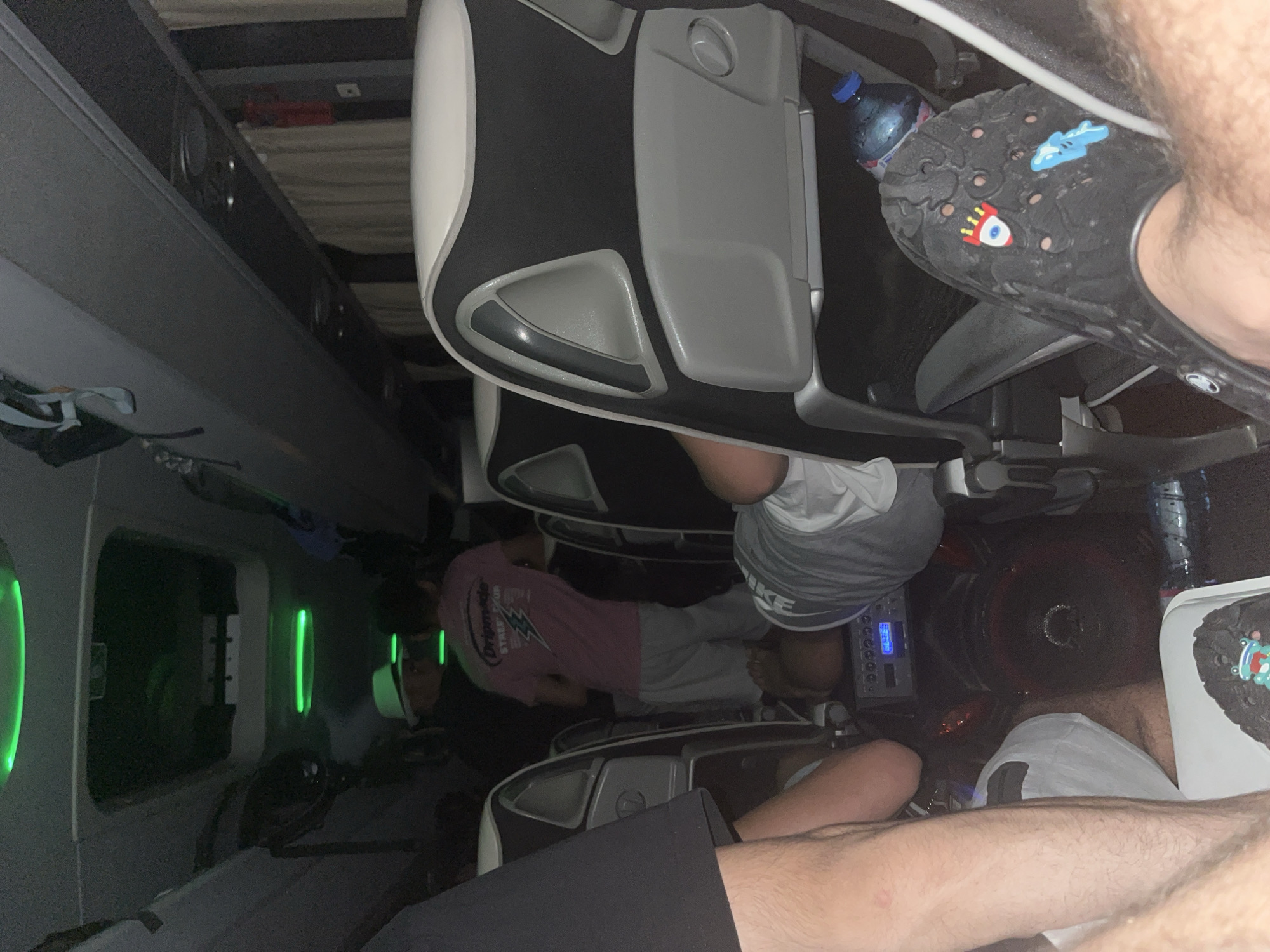 The image depicts the interior of what appears to be a sleeper bus, with rows of airplane-style seats. Passengers are seen seated, with some reclining. One person is wearing black Crocs with charms. A water bottle is visible near the seat. The presence of green lights suggests an entertainment or lighting system within the bus. There's no verifiable evidence suggesting the location is Slavovitsa, Bulgaria.