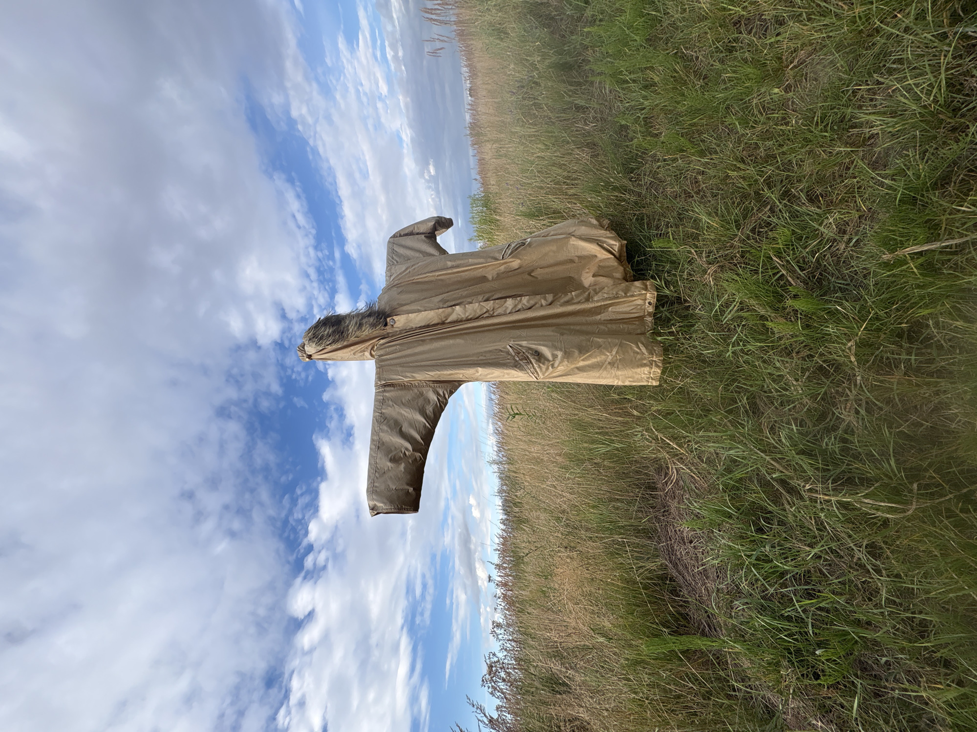 A solitary scarecrow, dressed in a tan hooded coat with fur trim, stands in a grassy field under a partly cloudy sky. The scarecrow is positioned in the center of the frame, its arms outstretched to the sides, resembling a crucifix. The field is overgrown with tall grasses and some wildflowers. In the background, more tall grass and distant trees are visible. The sky is a mix of blue and white clouds, with sunlight filtering through. The location is identified as Wilczyce, Poland. There are no people or animals present in the image. The scene appears peaceful and undisturbed, with the scarecrow serving as the sole focal point.