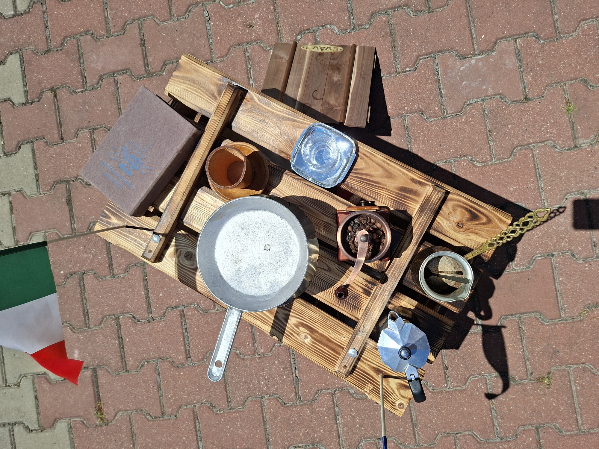 A wooden tray with various items is placed on a red brick paved ground. The tray has a rustic design with visible wood grain and some charring. On the tray, there is a silver-colored frying pan, a small copper coffee grinder with coffee beans, a silver moka pot, a small metal pitcher with a handle and a lid, and a small wooden box. To the left of the tray, an Italian flag is planted in a small hole. To the right of the tray, there is a book with a dark cover and blue writing on it, a small wooden cup with a saucer, and a small square glass dish. The scene is outdoors, under sunlight, casting shadows on the ground. There are no people visible in the image. The location is identified as Șerpeni, Moldova.