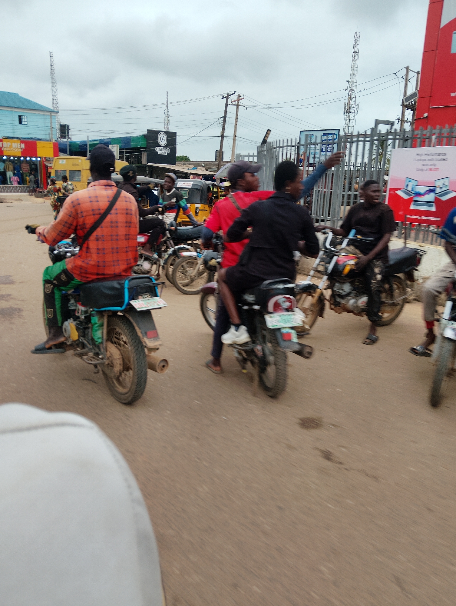 A group of people are riding motorcycles on a street in Agege, Nigeria. The sky is overcast with clouds. In the background, there are buildings, including one with a blue roof and a store called "TOP MEN BOUTIQUE". There are also several cellular towers visible. The road is made of asphalt. The people on the motorcycles are wearing various clothing, including a person in an orange and black plaid shirt, another in a red shirt, and one in a black shirt. Some of the motorcycles have passengers. There is also a yellow van and what appears to be a tricycle in the background. A billboard with advertisements is visible behind a metal fence on the right side of the image. The image appears to be taken from a moving vehicle.
