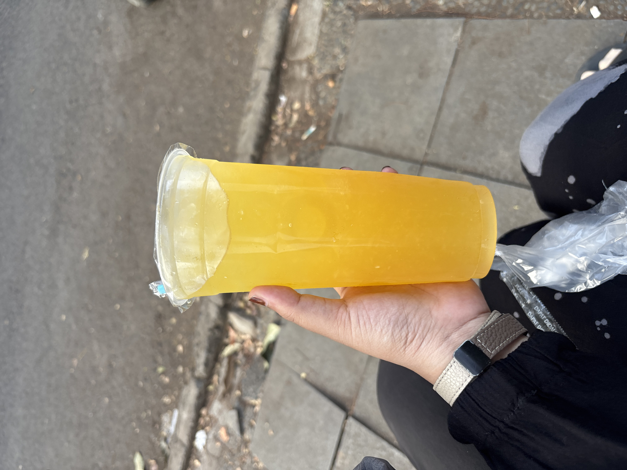 A person's hand is holding a large, clear plastic cup filled with a bright yellow beverage. The cup has a clear plastic lid, and a small blue tab is visible on the lid. The beverage appears to be cold, with condensation on the outside of the cup and small bubbles within. The person is wearing a black garment and a light-colored watch with a textured strap. Below the cup, a clear plastic bag is visible, containing two straws. The background shows a sidewalk made of grey concrete pavers, with a road surface to the left. Debris and some dried leaves are scattered on the ground. The overall scene suggests an outdoor setting, likely in a public area.