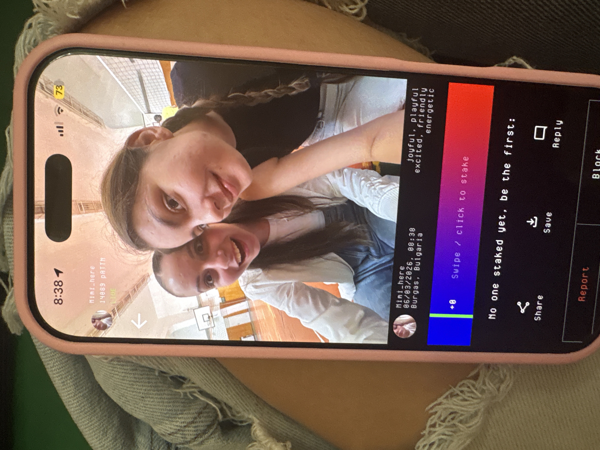The image displays a smartphone, encased in a pink cover, viewed from an overhead, rotated perspective, revealing its screen content. The phone's screen shows a selfie of two young women smiling broadly at the camera. Their expressions convey happiness and playfulness. The background of their selfie appears to be an indoor sports facility, with a white net or barrier and a basketball hoop's backboard visible.
The phone's interface is active, likely from a social media or content-sharing application. The time displayed on the phone is 8:38, with a battery icon showing 73% charge. Text on the screen identifies the user as "Mini_here" and specifies the post details: "06/03/2026, 08:38," and the location as "Burgas, Bulgaria." Associated with the post are descriptive words: "joyful, playful, excited, friendly, energetic."
Other interactive elements visible on the screen include a count of "+8," a prompt to "Swipe / click to stake," and the message "No one staked yet, be the first:". Below this are options to "Share," "Save," "Reply," "Block," and "Report." Another instance of the username "Mini_here" is visible with "14089 PATTN" and "LIVE" beneath it. The foreground of the overall photo shows textured fabric, possibly denim, where the phone rests.