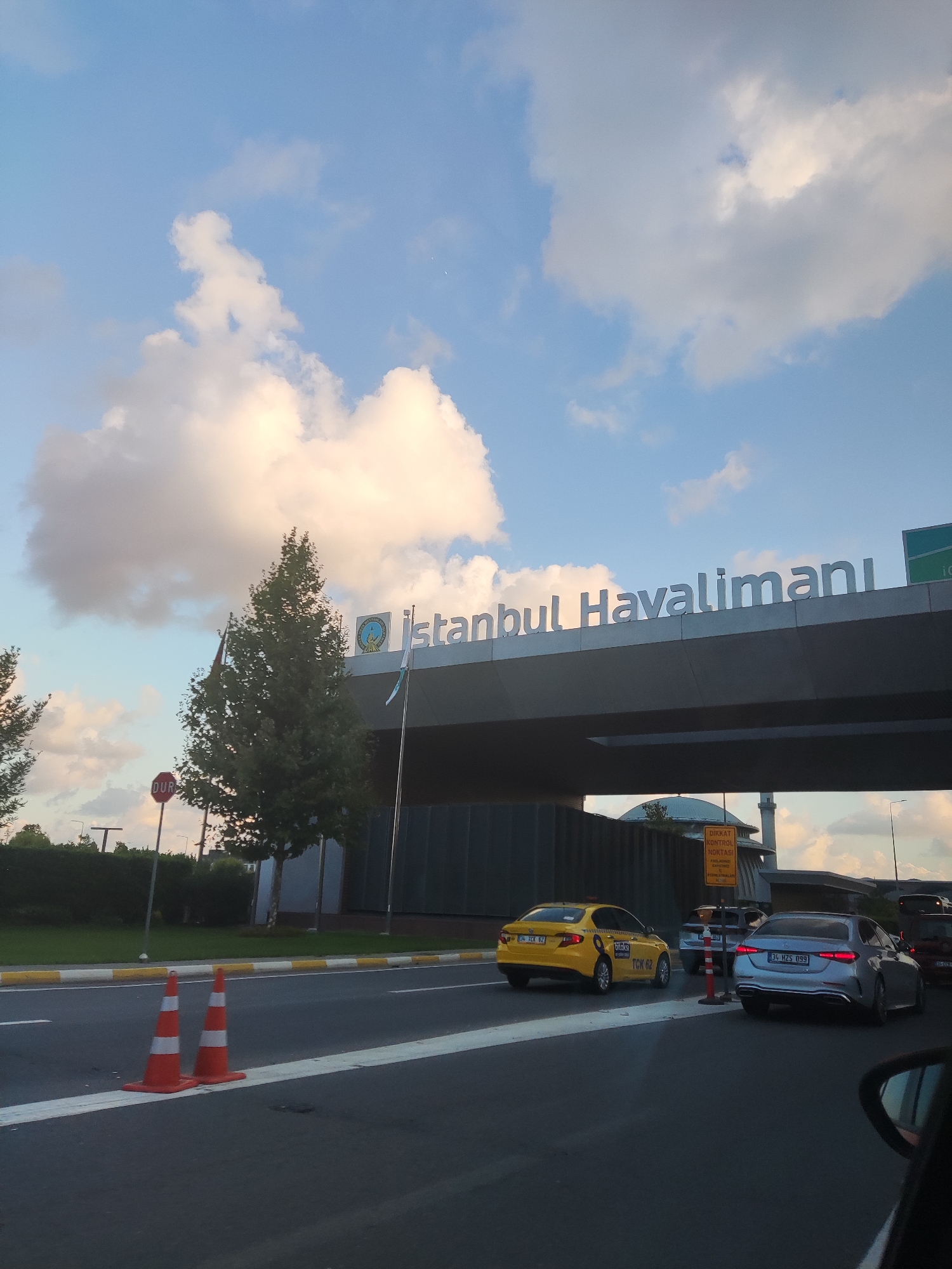 The image shows a view of the Istanbul Airport (İstanbul Havalimanı) in Arnavutköy, Turkey. The airport's name is prominently displayed in large white letters on an elevated structure. The sky above is partly cloudy with a light blue hue, and the sun appears to be setting or rising, casting a soft light.
In the foreground, there is a road with white and yellow lane markings. Two orange traffic cones with white reflective stripes are placed on the road. Several cars are visible on the road, including a yellow taxi with "iTaksi" branding and the license plate "34 TCK 62". A silver Mercedes-Benz sedan with the license plate "34 HZS 099" is also in view.
To the left, there is a red stop sign that says "DUR" (which means "STOP" in Turkish). A tree with green foliage stands near the stop sign, and beyond that, there are more trees and greenery. A dark-colored building, possibly part of the airport infrastructure, is visible in the middle ground. In the background, a mosque with a dome and a minaret can be seen, along with a yellow sign that reads "DİKKAT KONTROL NOKTASI" (which means "CAUTION CONTROL POINT"). The overall scene depicts a typical view of an airport entrance.