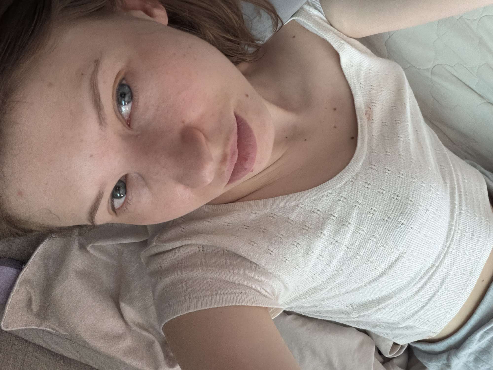 A young Caucasian female with light skin and freckles is lying down, looking up at the camera. She has blue eyes, light brown hair, and is wearing a white knitted tank top and grey pajama pants. The background consists of bedding in light beige and grey tones. The scene appears to be indoors, likely in a bedroom.