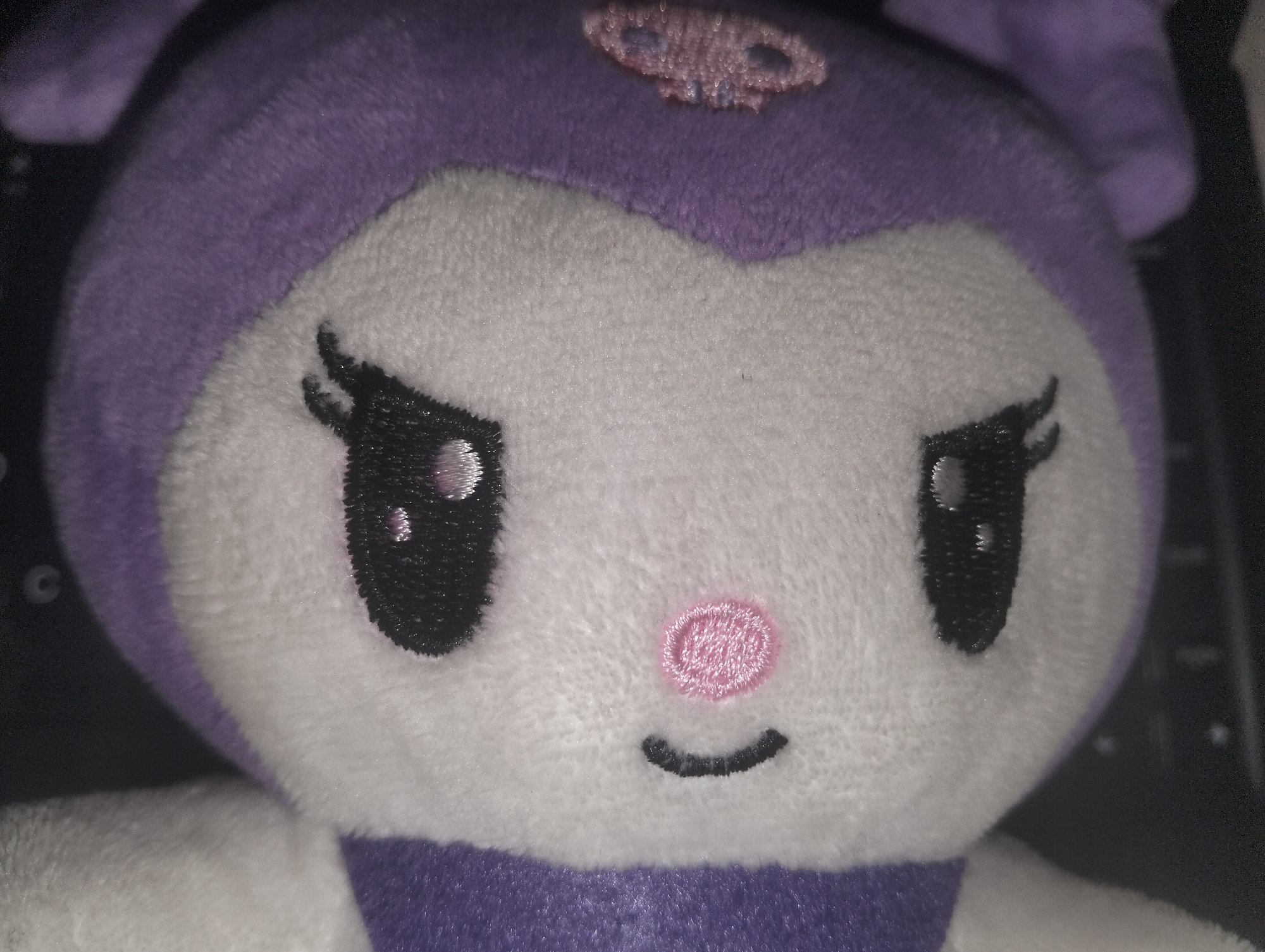 A close-up of a Kuromi plush toy shows its face. The plush is primarily purple and white with embroidered details: black eyes with white highlights, a pink, rounded nose, and a curved black mouth. A purple skull emblem is centered on the forehead. The background is dark, indicating a possible indoor setting, with hints of what might be a keyboard visible in the periphery. This information does not provide any indication of a location in Dhaka, Bangladesh.