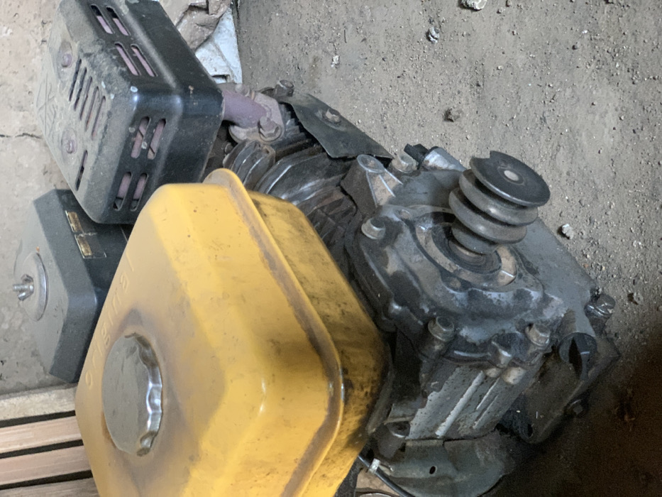 The image shows a close-up view of a small gasoline engine. The engine is a black, grey, and yellow color, and it is lying on its side on a concrete surface. The engine has a large yellow gas tank with a black cap on the top. The engine is located in the city of Egbeda, Nigeria.