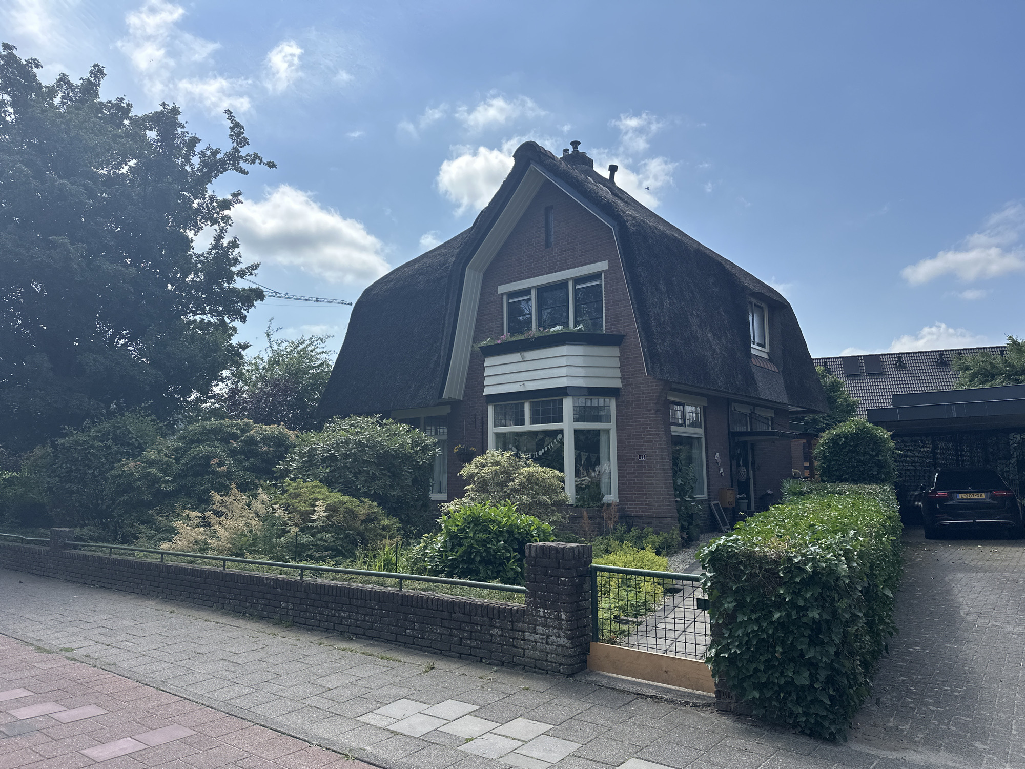 The image shows a brick house with a thatched roof and a large, well-maintained garden. A paved walkway leads to the front door, and a hedge lines the edge of the property. A car is parked in a driveway next to the house. The sky is blue with scattered clouds. The house is located in Steenwijk, Netherlands. There are no people or animals visible in the image.