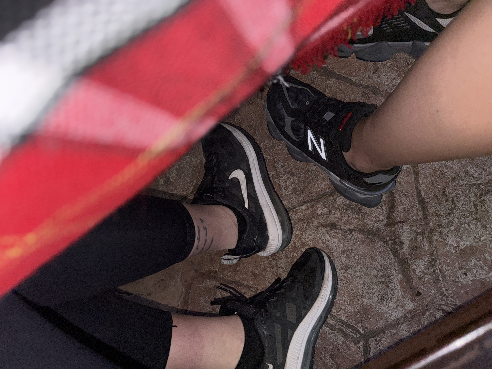 The image shows the lower legs and feet of two people sitting at a table. The table is covered with a red and white checkered tablecloth. One person is wearing black leggings and black Nike sneakers with white soles and a white swoosh. The other person is wearing black New Balance sneakers with a white "N" logo. The floor appears to be tiled, possibly brick or stone. The location is not identifiable as Sofia, Bulgaria from visual clues in the image.