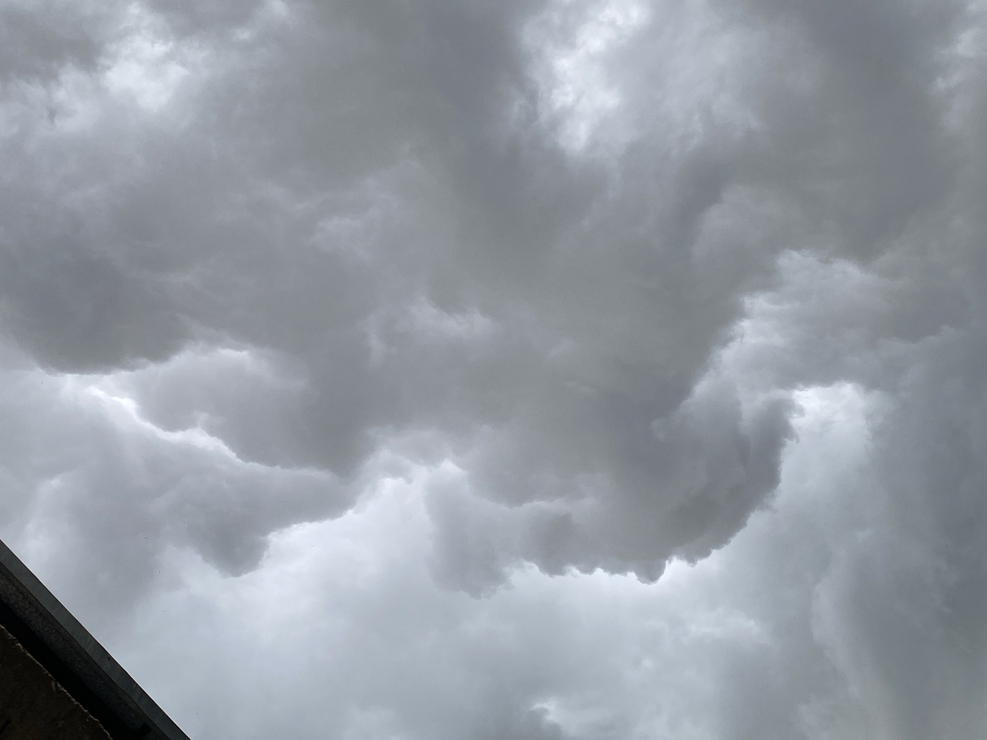 A low-angle view captures a sky filled with ominous, dark grey storm clouds. The clouds are dense and turbulent, suggesting an impending weather event. In the upper left corner, a portion of a dark, textured surface, possibly the underside of a roof or overhang, juts into the frame, providing a stark contrast to the vast expanse of cloud. No people or specific landmarks are visible in this image, which focuses solely on the atmospheric conditions. The location is identified as Uchaly, Russia.