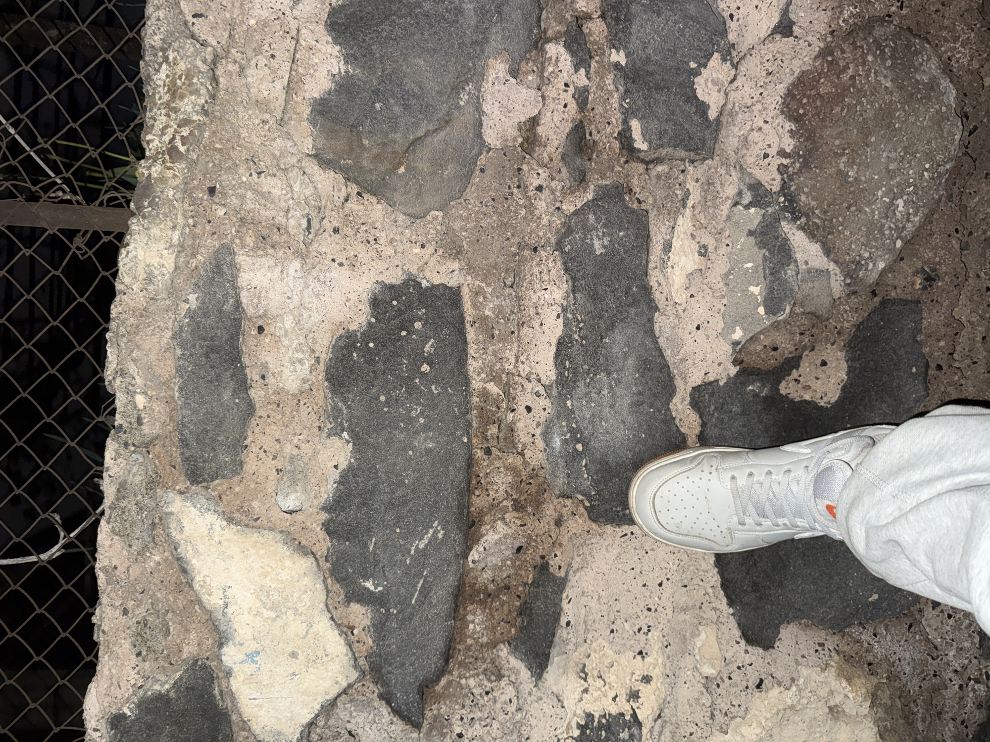 The image captures a top-down view of a person's right foot resting on a textured stone surface, likely in Yerevan, Armenia. The foot is clad in a light grey or white sneaker with white laces and an orange interior lining, paired with light grey sweatpants. The ground or wall consists of large, irregular dark grey stones embedded in a lighter grey or beige mortar, which shows signs of wear and a rough texture. Some areas of the mortar appear chipped or eroded. On the far left of the frame, a dark chain-link fence is visible, suggesting an outdoor setting. The background behind the fence is dark, implying that the image was taken at night, with artificial lighting illuminating the scene. No specific activity beyond standing is depicted, and no text is visible.