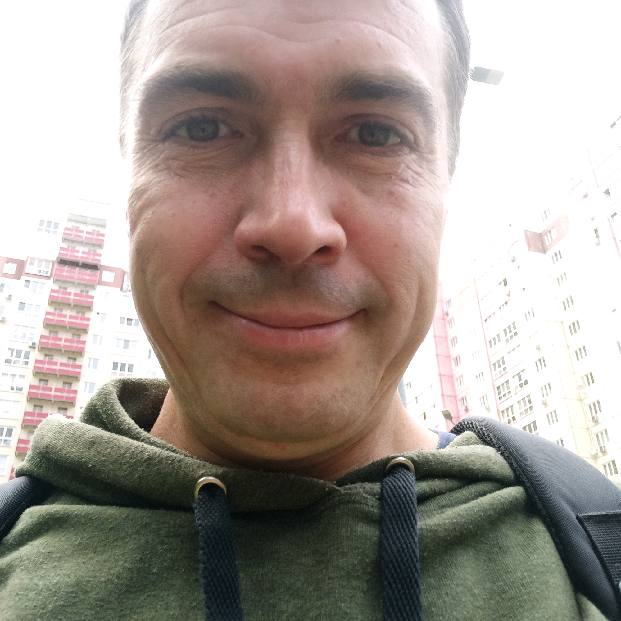 A close-up selfie of a man wearing a green hoodie is taken outdoors. The man's face is centered in the frame, with the background showing a tall apartment building with pink balconies and numerous windows. The sky is bright and slightly overcast. To the right of the man's head, a street lamp is visible. The man appears to be smiling slightly. The lower portion of the image shows the green hoodie and the straps of a backpack. The overall scene suggests an urban environment in Nizhny Novgorod, Russia, likely taken during the daytime.